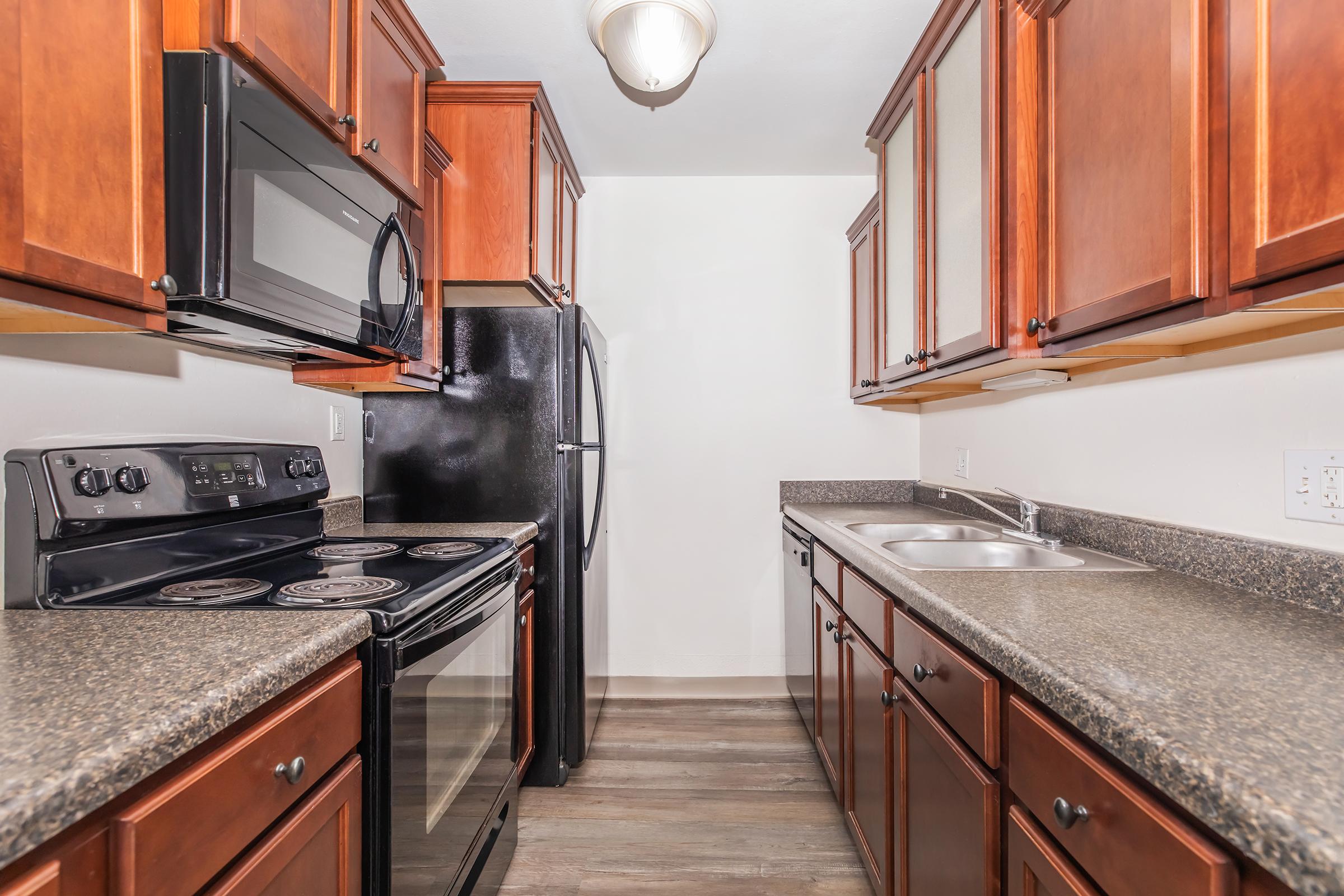 A modern kitchen featuring wooden cabinetry, granite countertops, and stainless steel appliances. It includes a black oven, stove, and microwave, along with a black refrigerator. The kitchen layout is compact with a double sink and ample storage, highlighting a clean and contemporary design.
