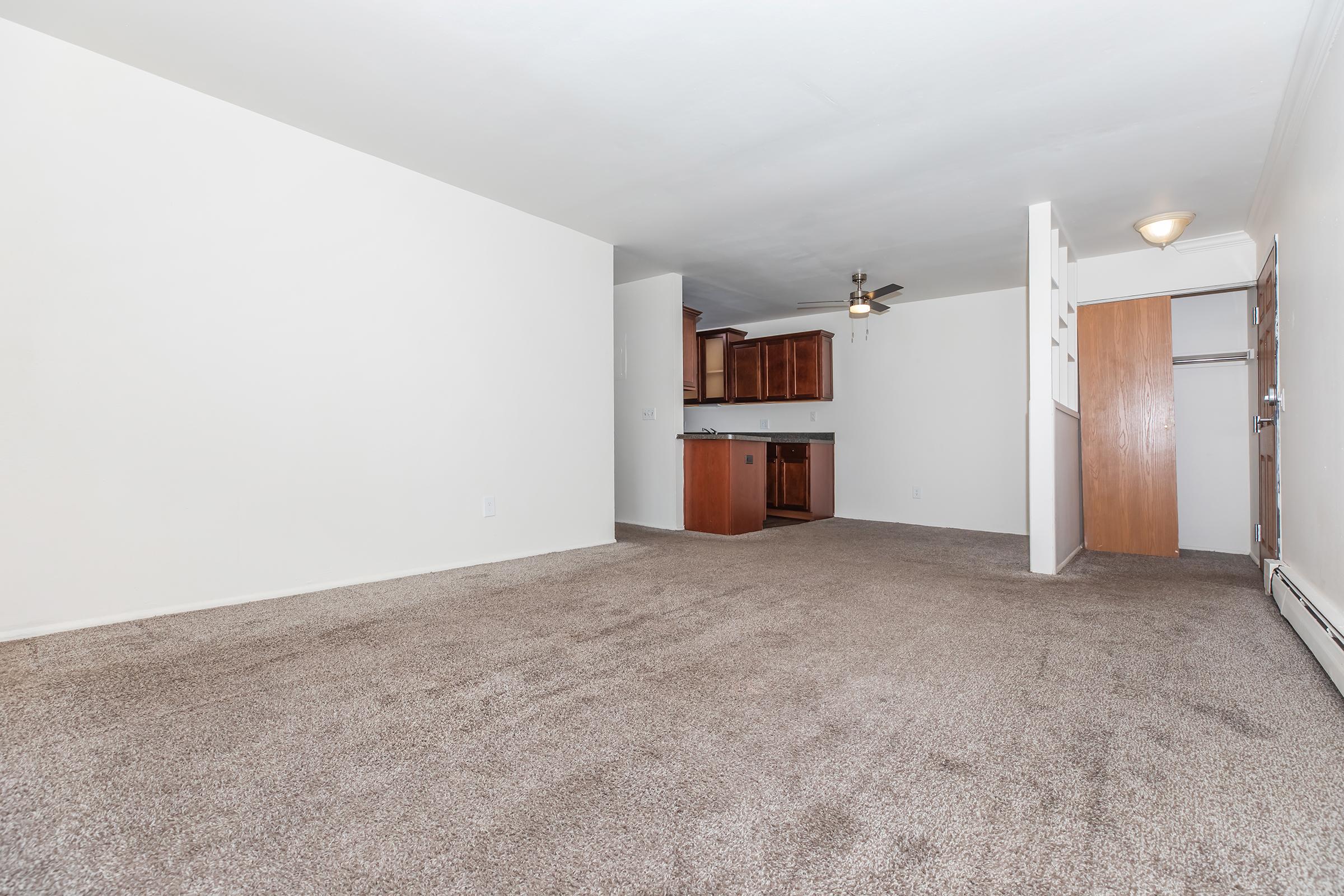 A spacious, empty living room with light-colored walls and a tan carpet. In the background, there is a doorway leading to a kitchen area featuring wooden cabinets. A ceiling fan is visible, and the room is well-lit with natural light.