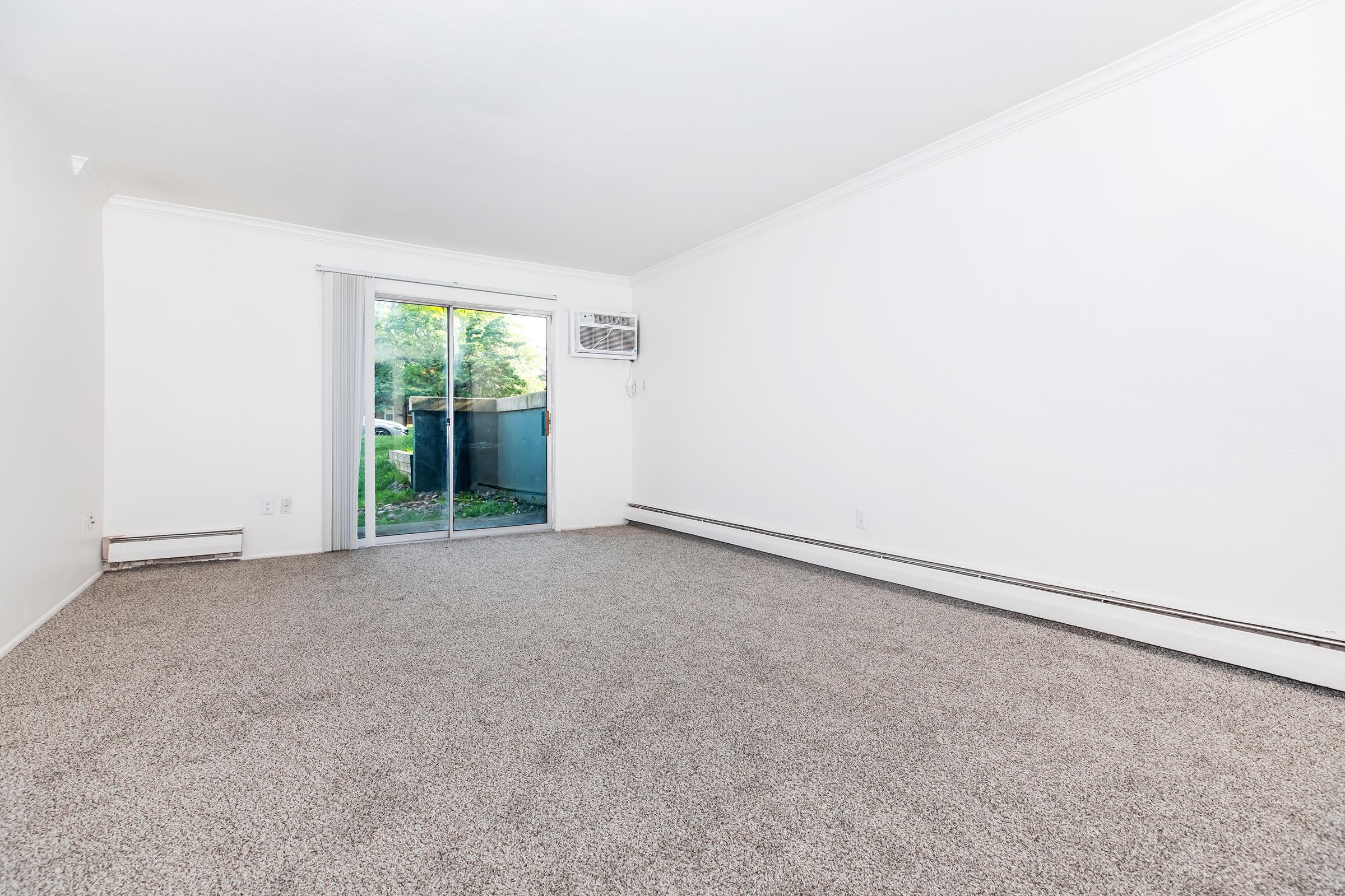 Empty living room with light beige carpet and white walls. A sliding glass door leads to an outdoor area, providing natural light. An air conditioning unit is mounted on the wall near the door. The space appears clean and well-maintained, ready for furniture placement.