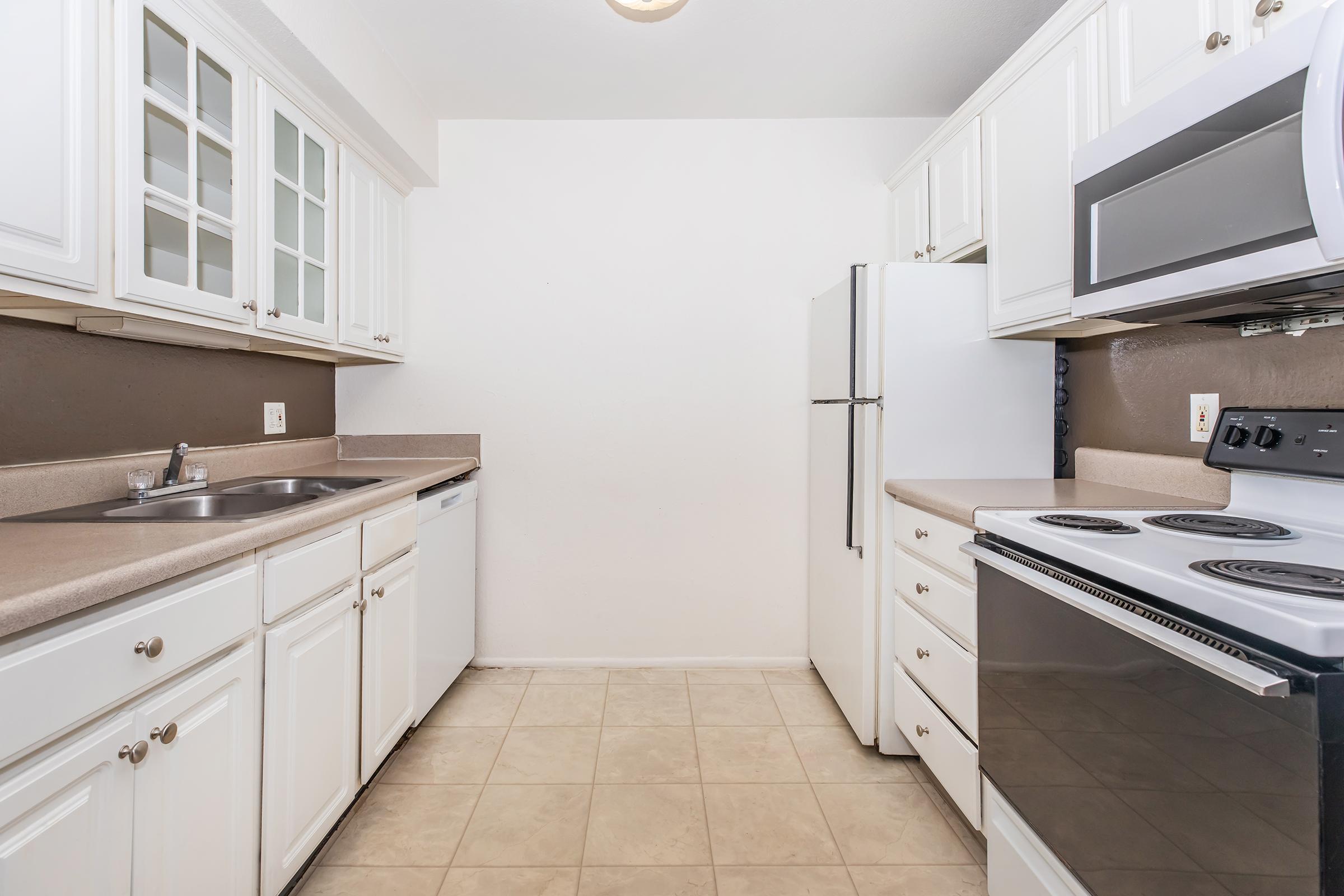 A modern kitchen featuring white cabinetry, beige countertops, and an open layout. Appliances include a refrigerator, microwave, stovetop, and oven. The walls are painted in a neutral tone, and the floor is tiled. Natural light enhances the space.