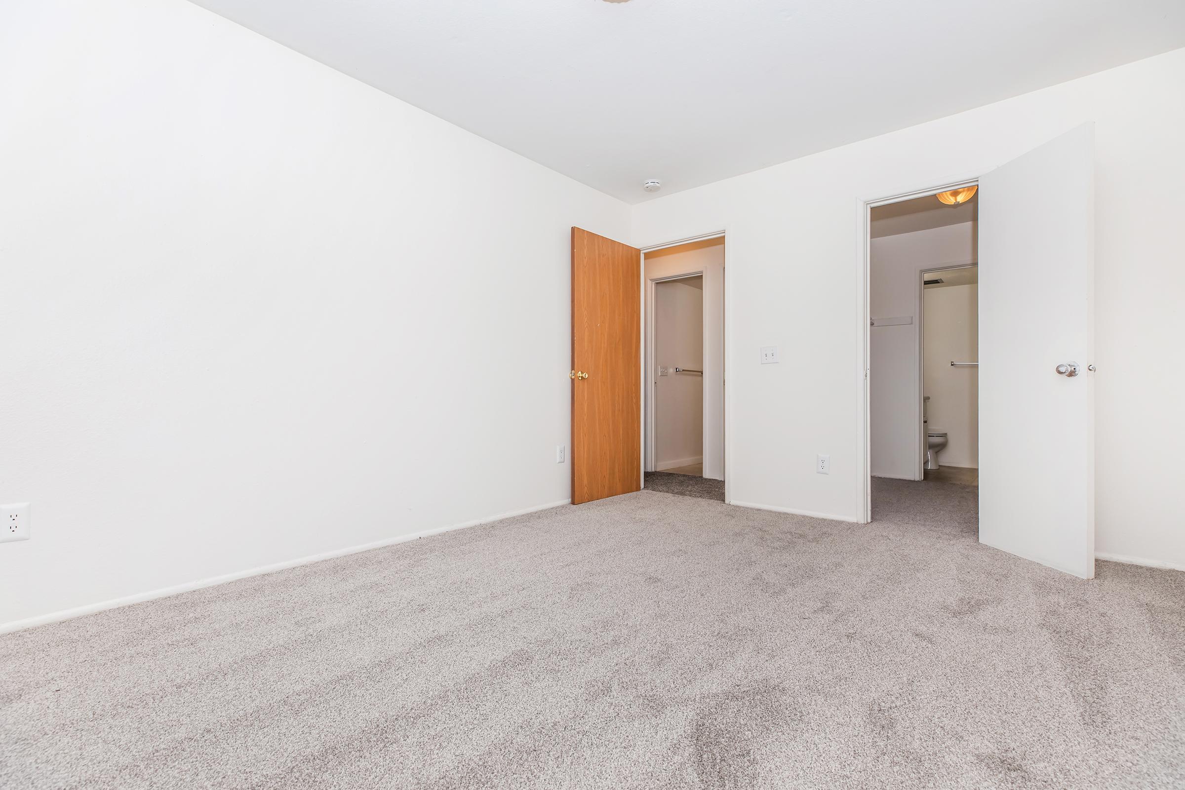 A light, empty room with beige carpet and white walls. Two doorways are visible: one leading to a small bathroom, the other to a hallway. Natural light enters from an open door, creating a spacious and clean atmosphere. Ideal for various uses or personal decoration.