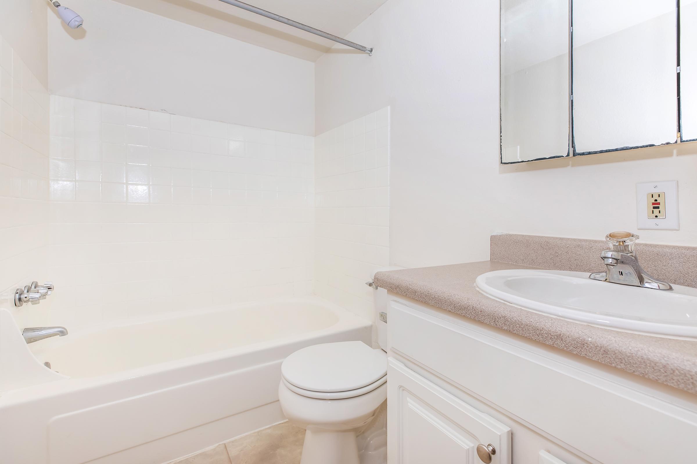 A clean and modern bathroom featuring a white bathtub, a toilet, and a sink with a countertop. The walls are tiled with white tiles, and there are mirrors above the sink. The overall appearance is bright and tidy, creating a fresh atmosphere.