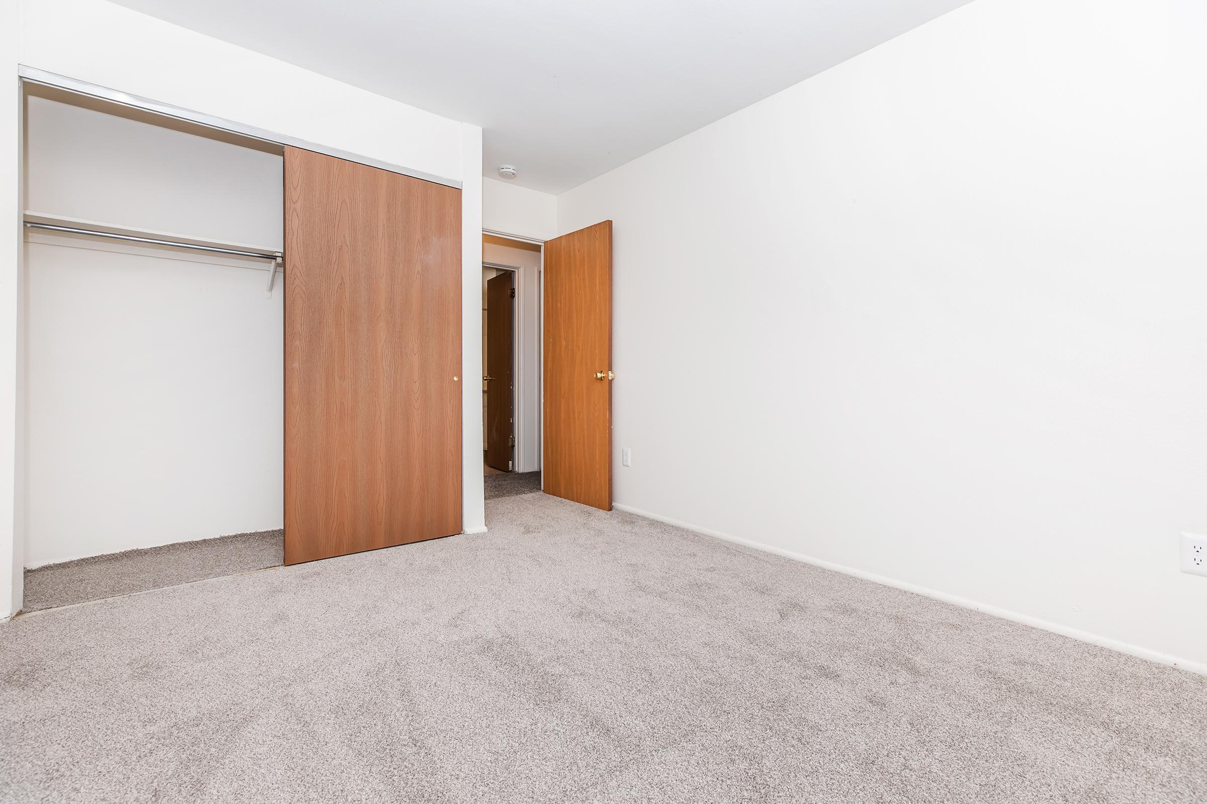 A spacious, empty room with light-colored walls and beige carpet. One corner features an open closet with sliding wooden doors, while a second door leads to another area. The room has a bright, inviting atmosphere, making it suitable for various purposes.