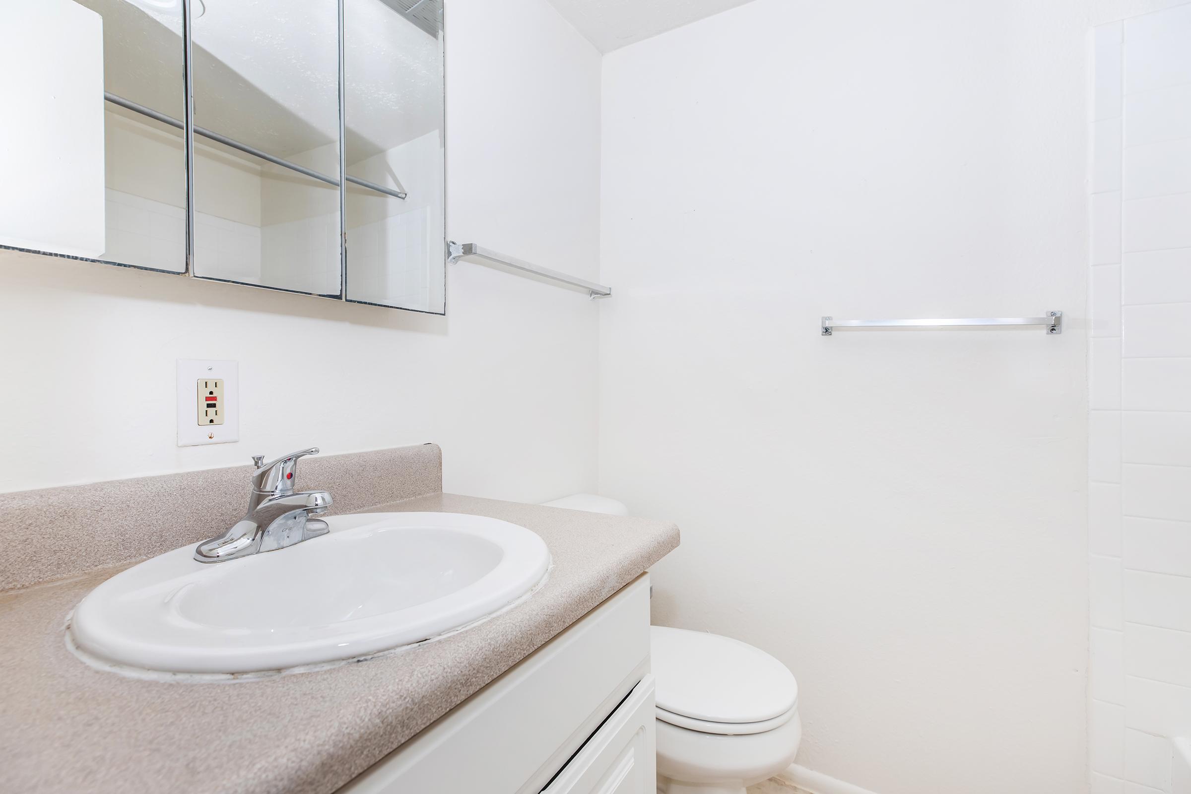 A clean bathroom featuring a white sink with a circular basin, a faucet, and a countertop. The walls are white, and there are mirrored cabinets above the sink. A toilet is positioned nearby, and there is a towel bar on the wall. The overall design is simple and modern, with ample natural light.