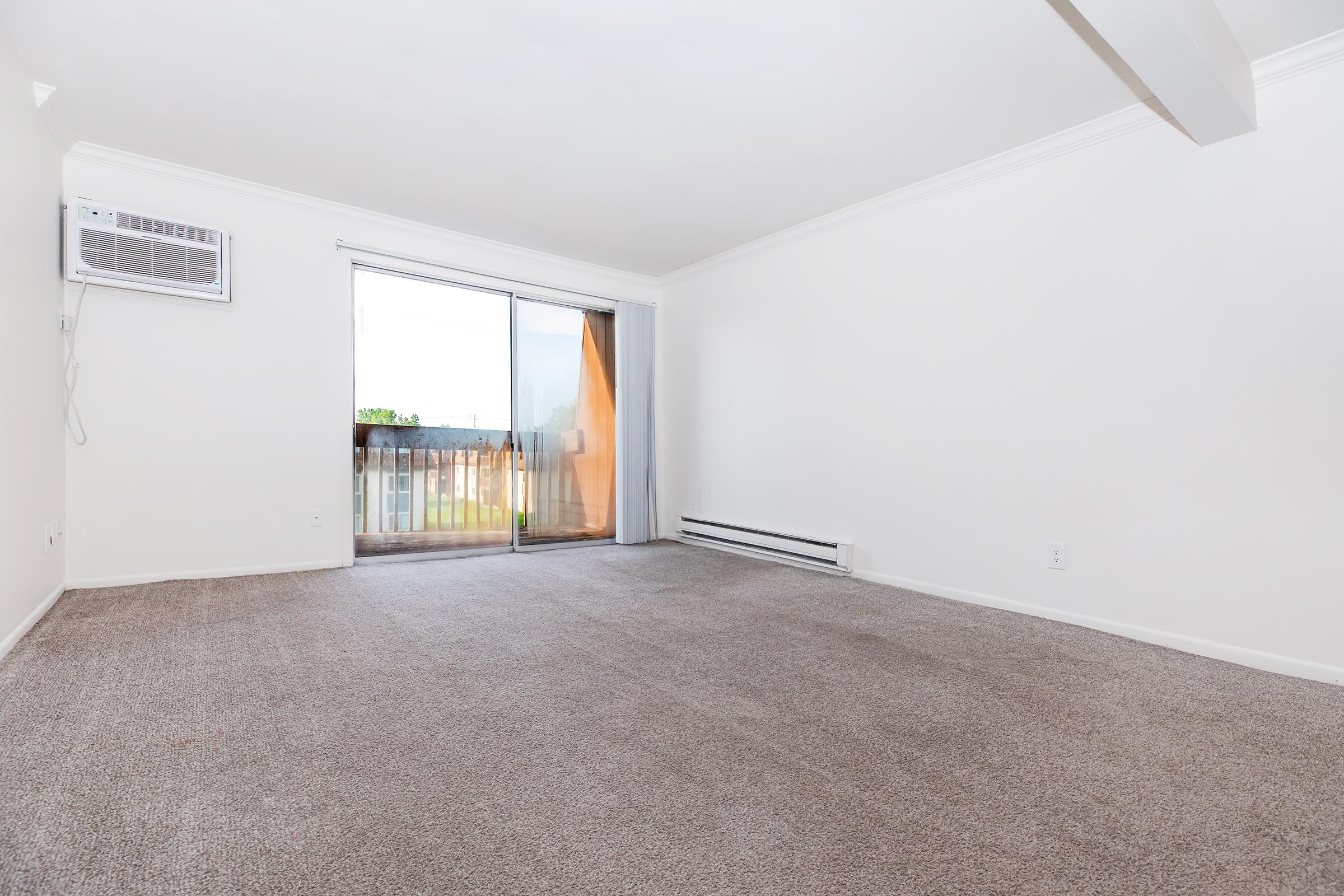 Empty room with light-colored walls and a large window covered by sheer curtains, allowing natural light to enter. The floor is carpeted in a neutral tone, and there is an air conditioning unit mounted on the wall. A sliding glass door leads to a small balcony area.