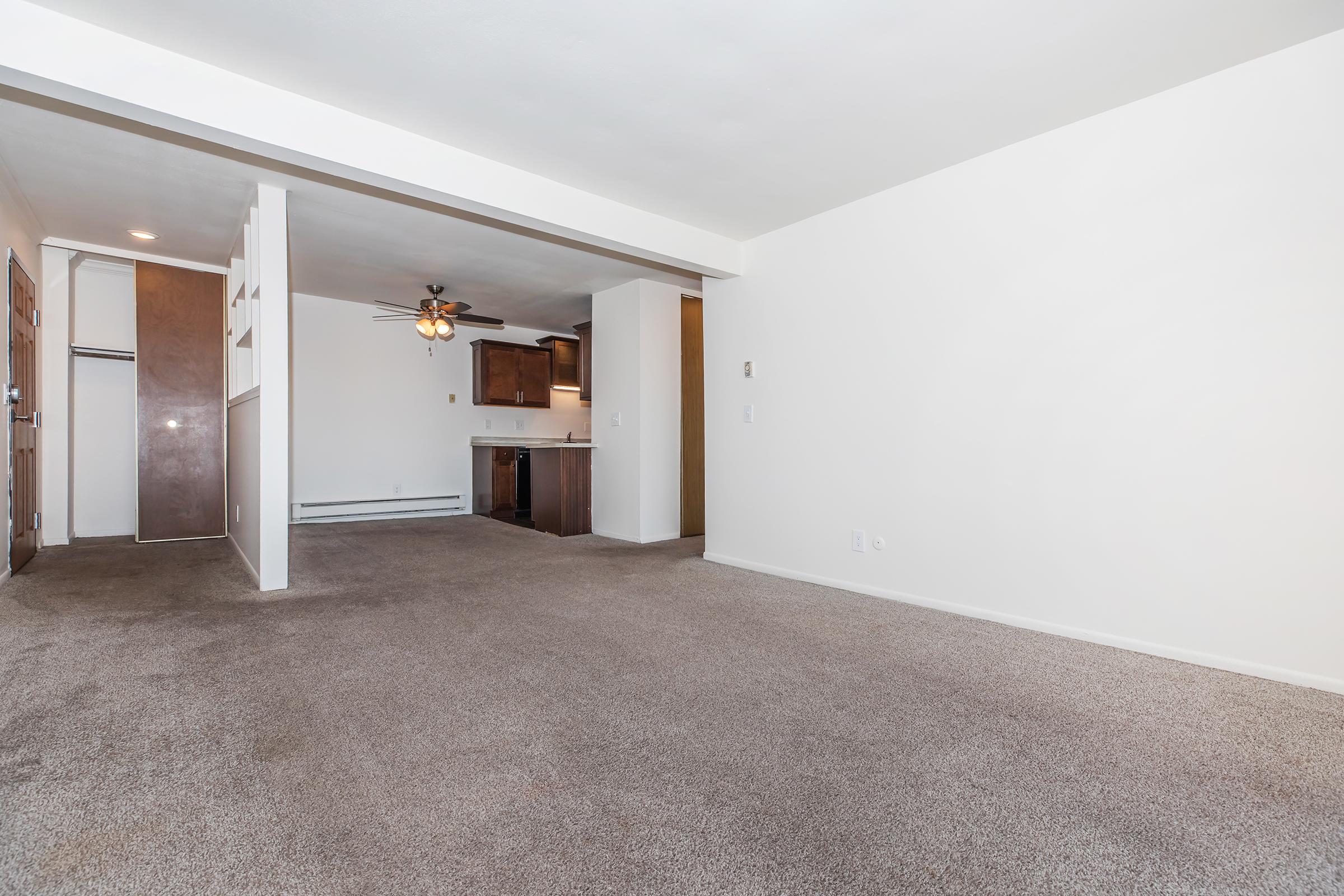 A spacious, empty living area with light beige carpet and off-white walls. To the left, a door leads to a closet, while a ceiling fan hangs above. Ahead is an open kitchen with dark wooden cabinets and a countertop. The overall atmosphere is bright and ready for furnishing.