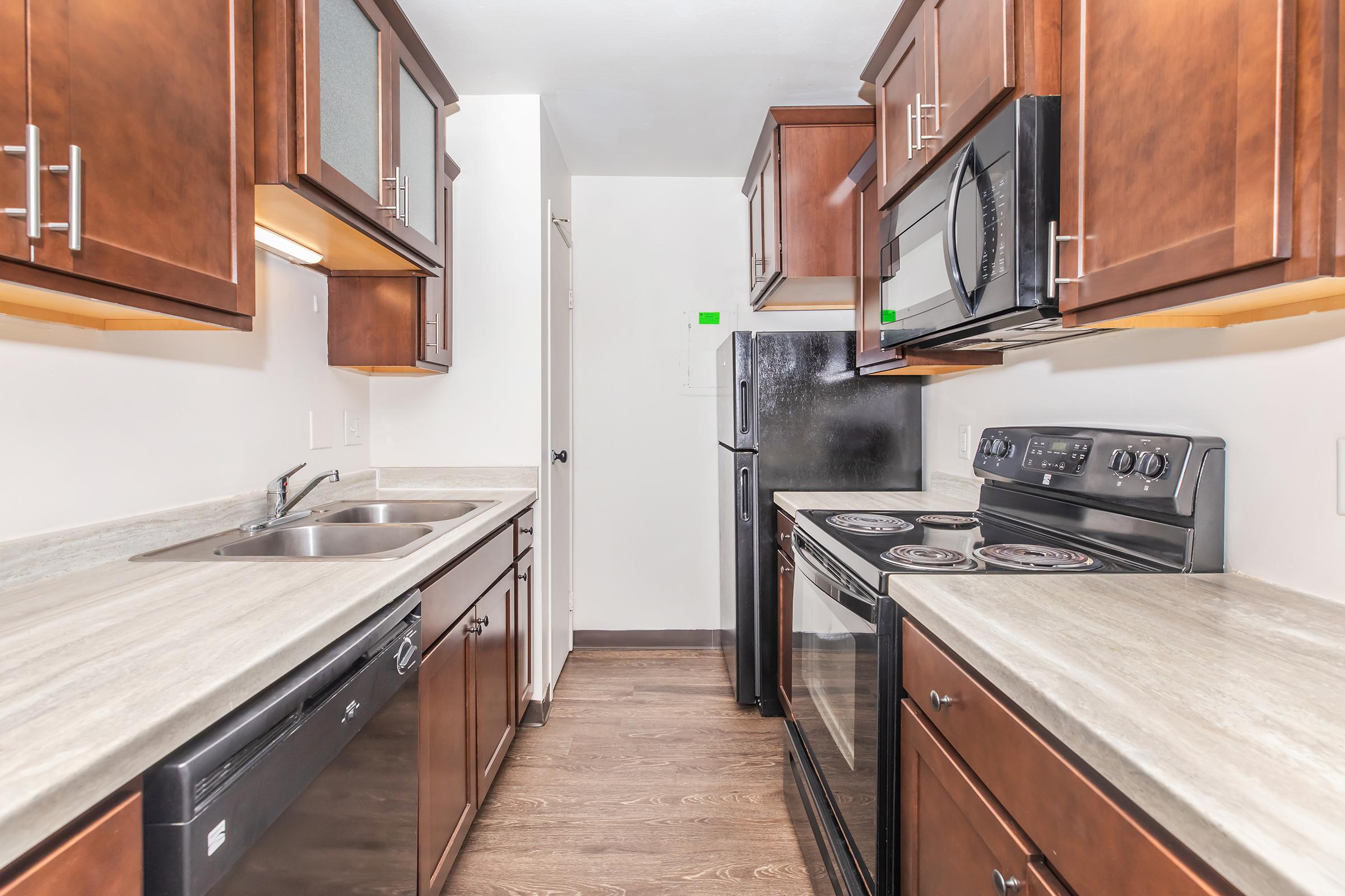 Modern kitchen featuring dark wood cabinetry, stainless steel appliances, and a grey countertop. The layout includes a double sink, a dishwasher, an electric stove, and a microwave, with a narrow corridor leading to a door at the end. The space is well-lit and neatly arranged.