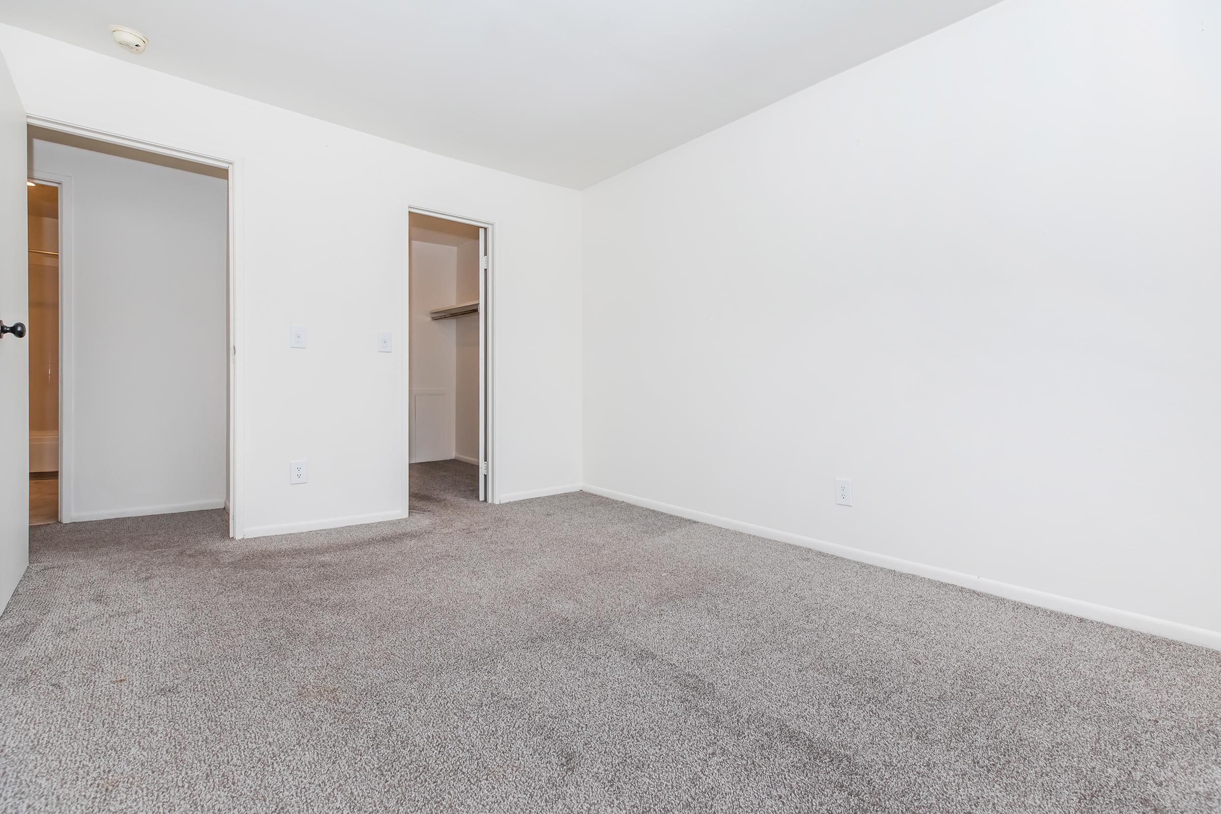 Empty room with beige carpet and white walls. Two doorways lead to other areas, one possibly a closet. Natural light comes from an unseen window, creating a bright and spacious atmosphere. The room is unfurnished, offering a blank canvas for decoration.