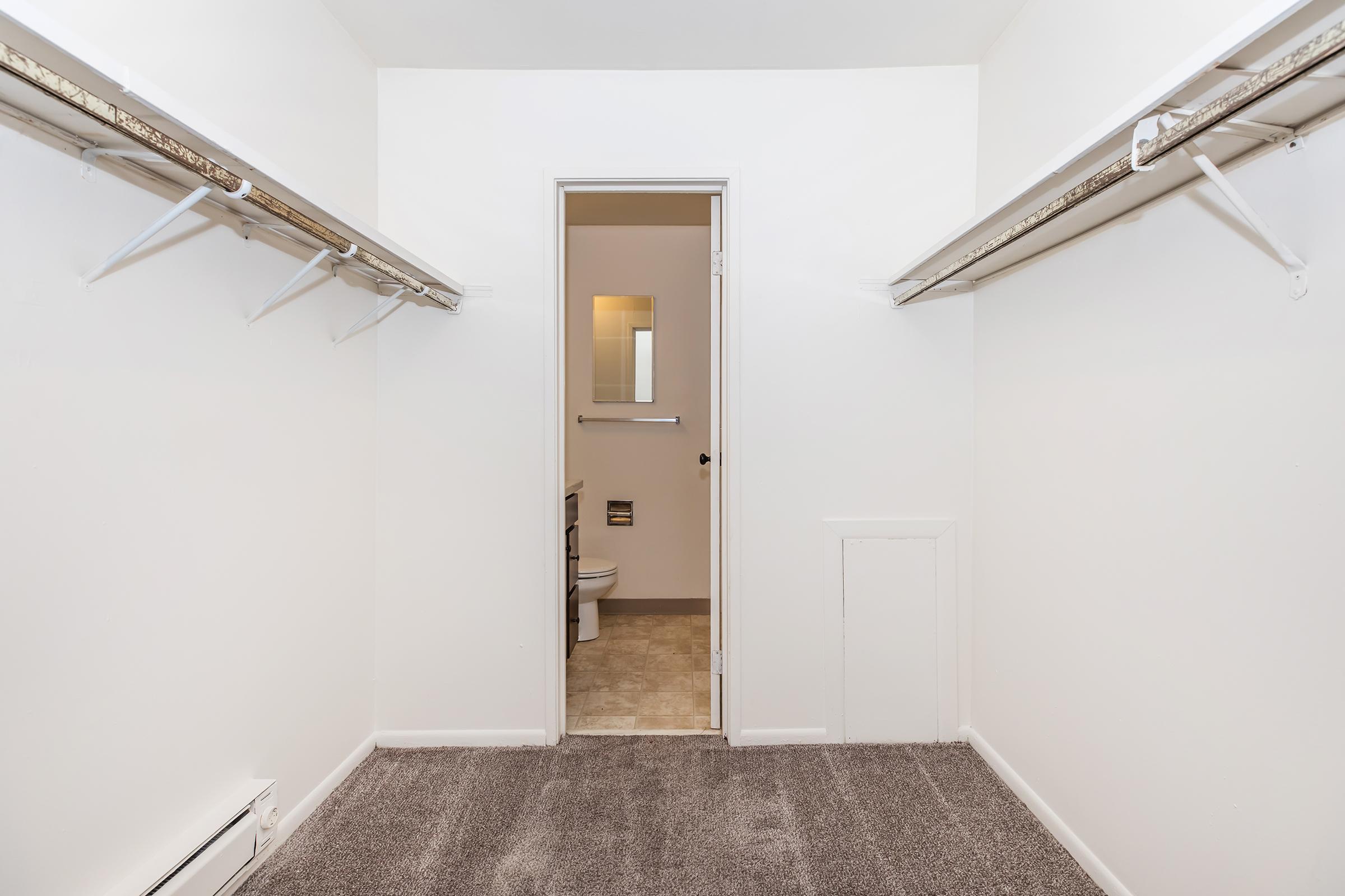 A spacious, empty closet with two hanging rods along the walls and carpeted flooring. A door at the far end leads to a small bathroom, which is partially visible. The walls are painted white, giving a clean and bright appearance to the space.
