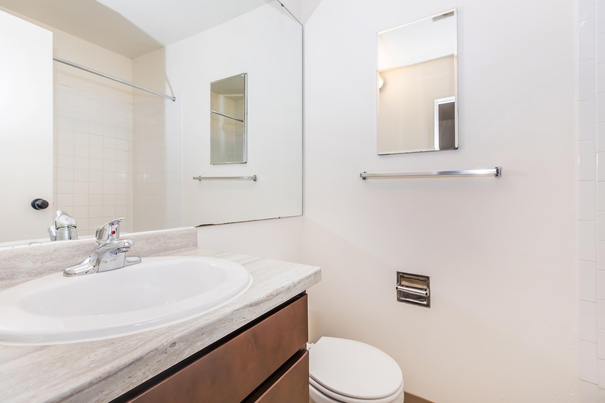 A clean bathroom featuring a sink with a sleek faucet, a small mirror above the sink, a wall-mounted towel bar, and a white toilet. The walls are painted light colors, and there is a tiled shower area visible in the background. The overall atmosphere is bright and minimalist.