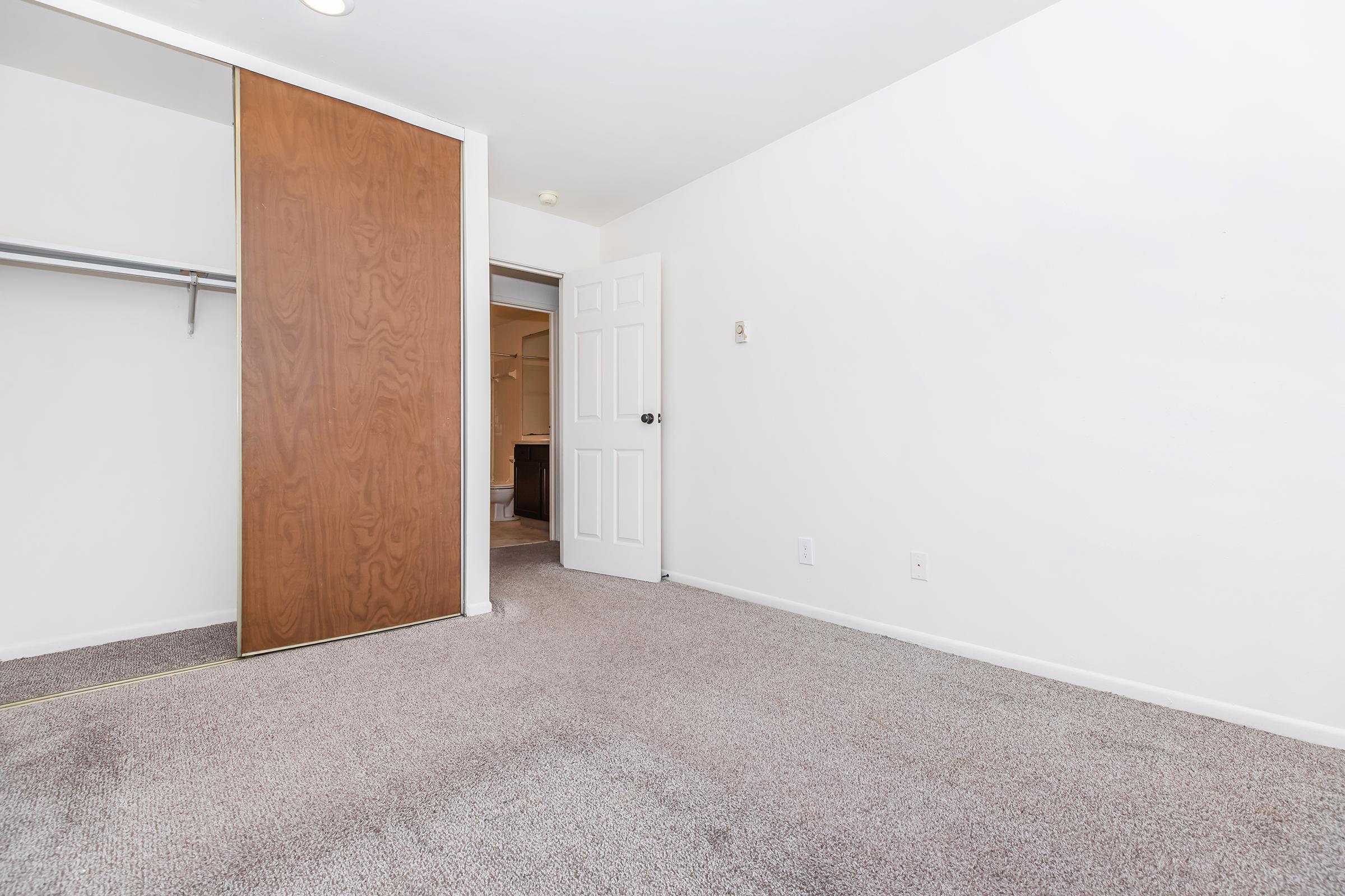 Empty room with light-colored walls and carpeted flooring. There's a closed wooden door on the left, leading to a closet, and an open door in the background leading to another room. The lighting is bright, and the space appears clean and uncluttered.