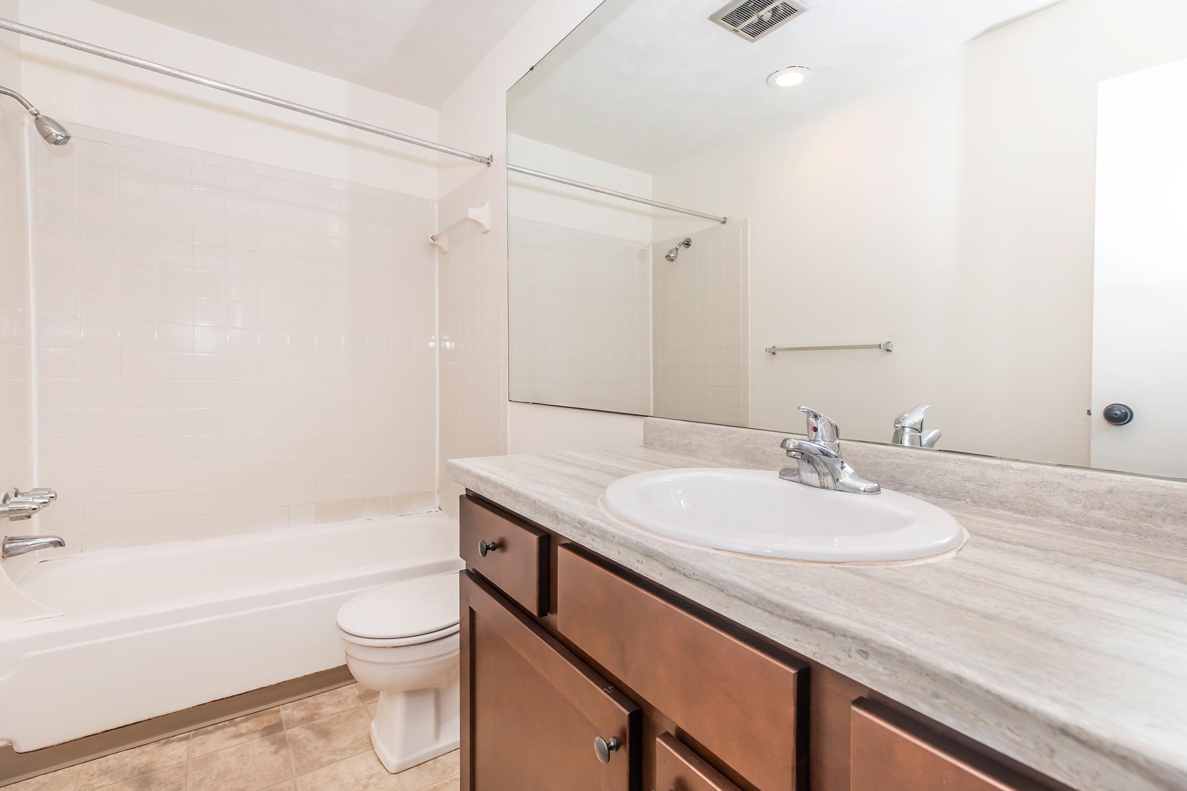 A modern bathroom featuring a white bathtub and toilet, with a large mirror above a vanity sink. The sink has a chrome faucet and the countertop is made of light-colored stone. The walls are painted in a neutral tone, and there is a towel bar mounted on the wall. Natural light brightens the space.