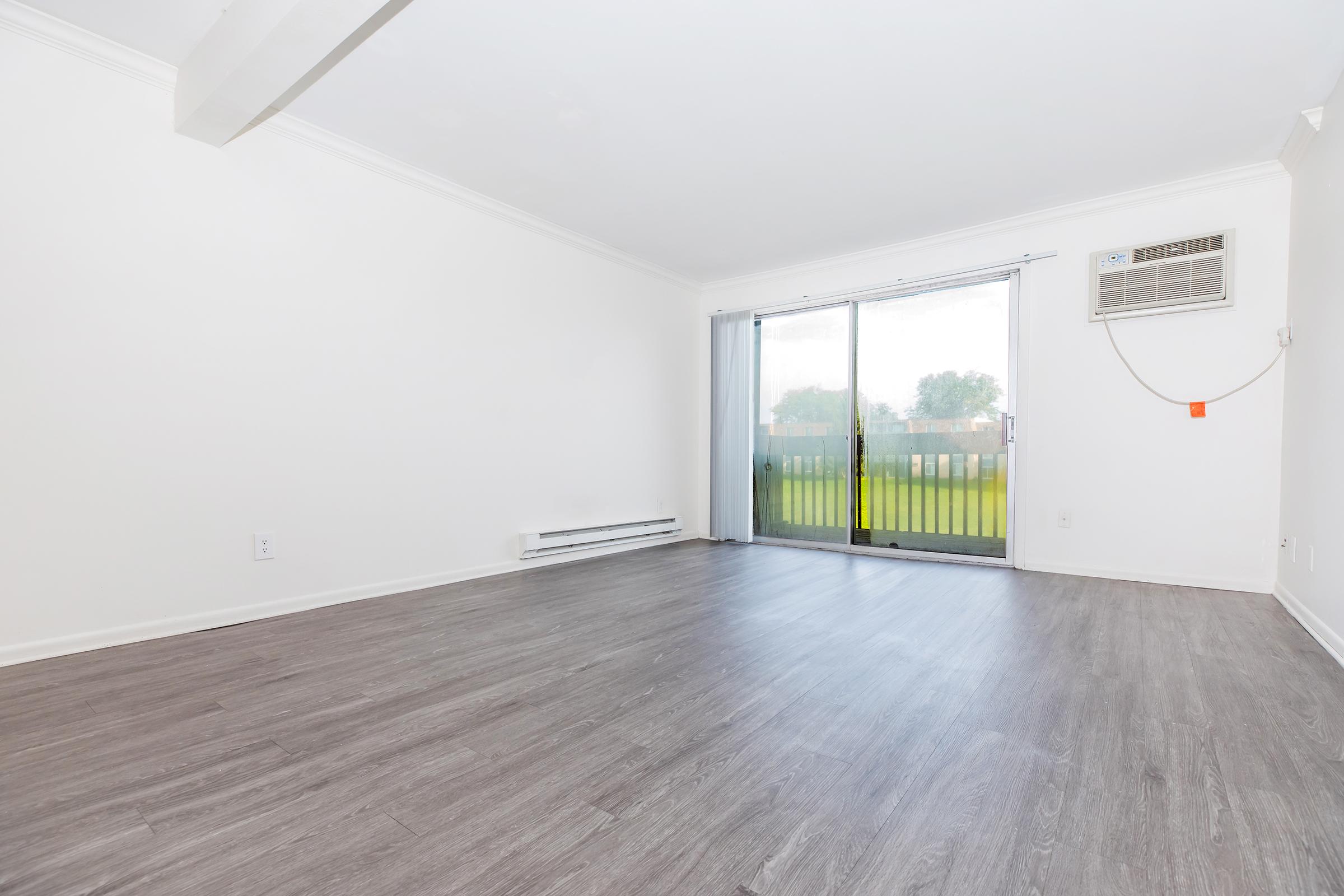 A spacious, empty room featuring light-colored walls and a large sliding glass door that leads to an outdoor area. The flooring is light grey and has a modern finish. An air conditioning unit is mounted on the wall, and there are no furnishings present, giving a clean, minimalist look.