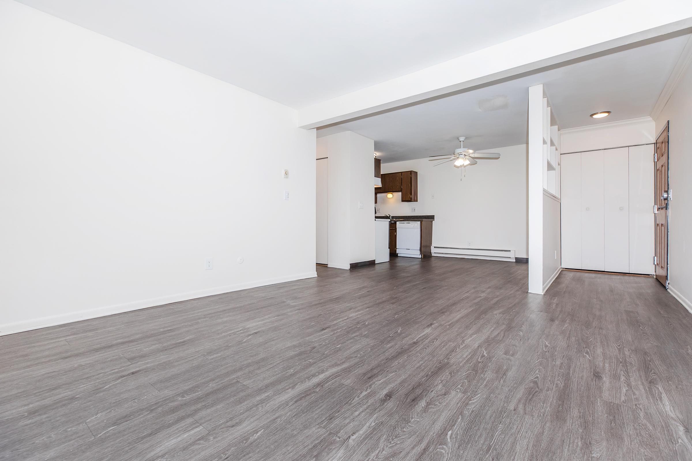 Spacious and modern living area with light-colored walls, a large window for natural light, and a view of a kitchen area with wooden cabinetry. The flooring is dark laminate, creating a contemporary look. The room features a ceiling fan and leads to an entrance with a door and closet.