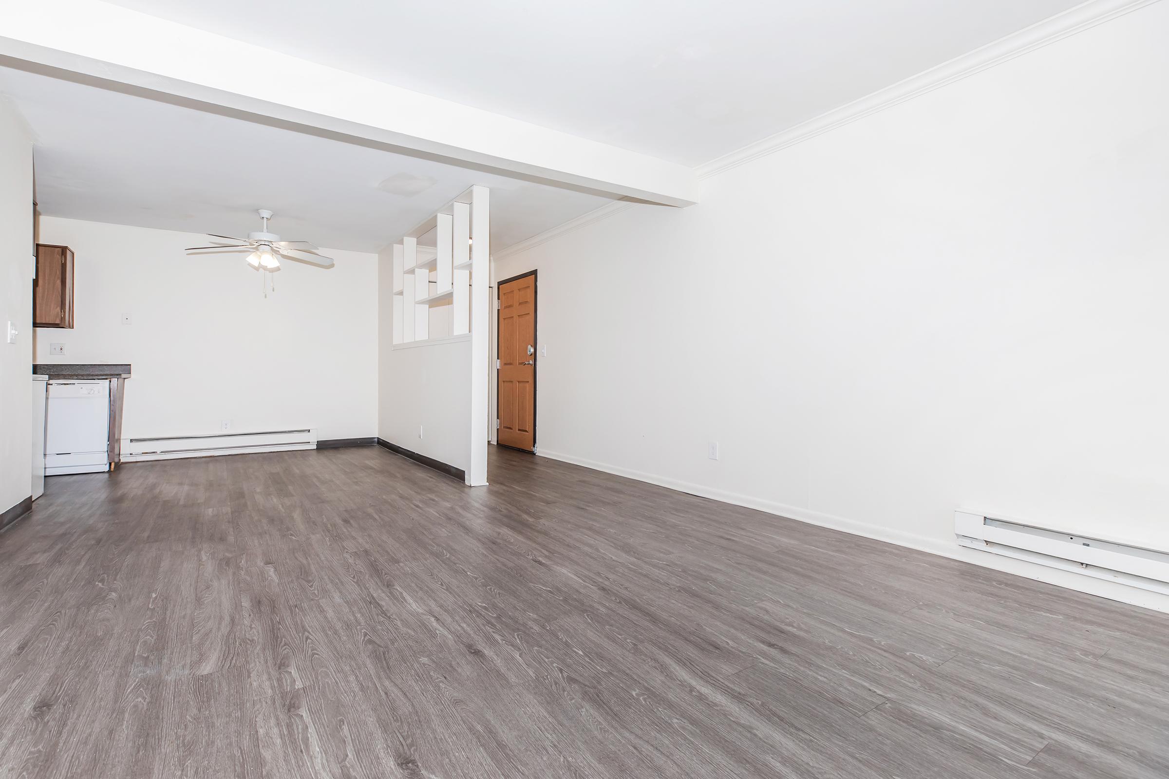 A spacious, unfurnished living area featuring light-colored walls and dark wood-like flooring. A ceiling fan is visible, and the room has a doorway leading to what appears to be an entrance. There is also a partial wall with an open design separating the living space from the kitchen area.
