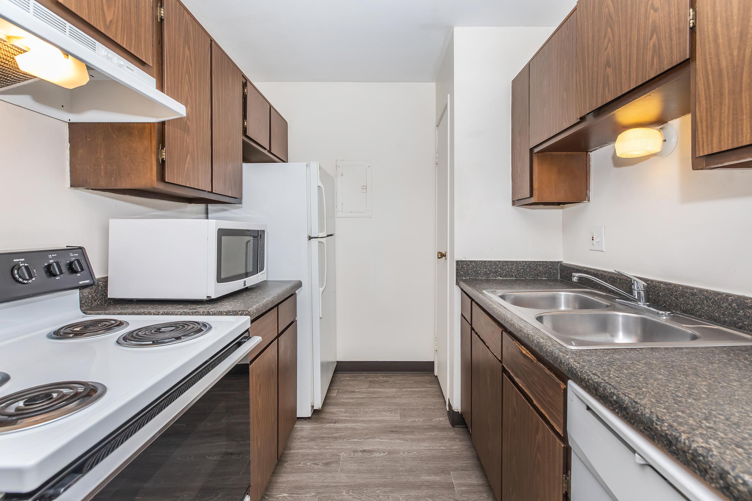 A compact kitchen featuring dark wooden cabinets, a black stove, and a microwave on the counter. There's a double sink with a granite countertop and a white refrigerator against the wall. The walls are painted a light color, and the flooring is a wood-like laminate.