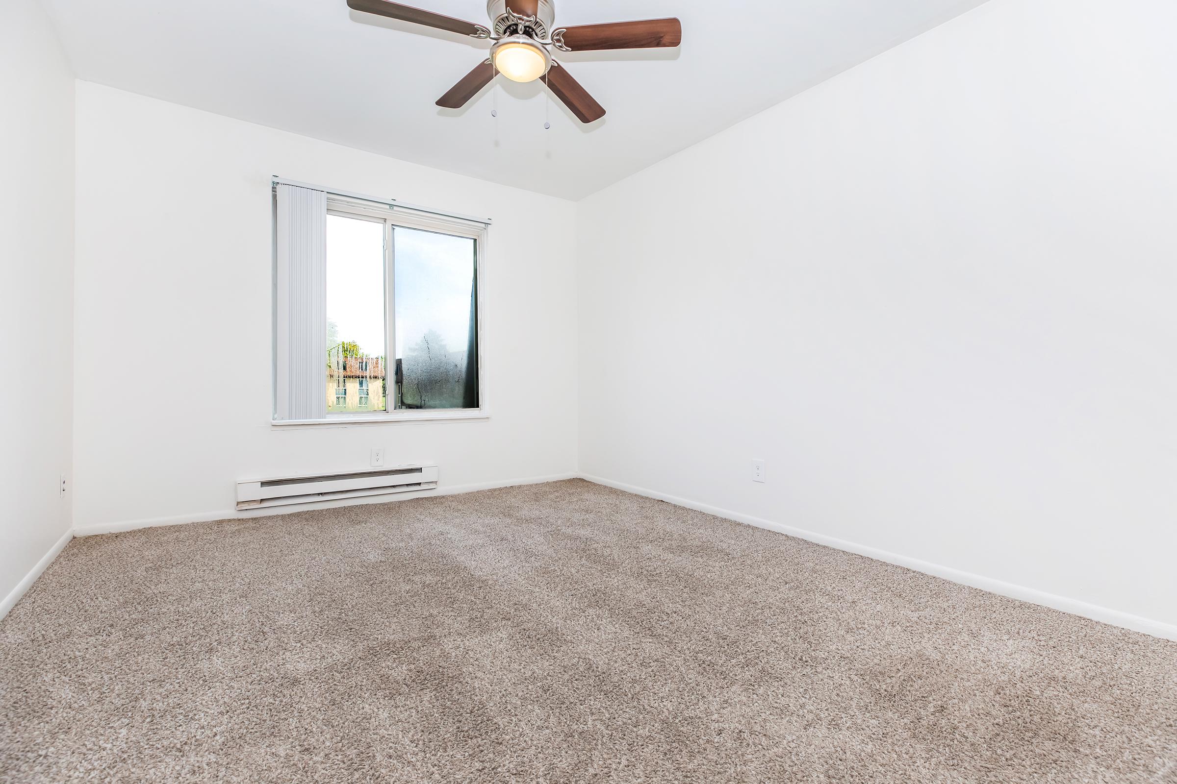 Empty room with beige carpet, a ceiling fan, and a large window allowing natural light. The walls are painted white, and there are no furnishings or decorations visible. A heating unit is located beneath the window.
