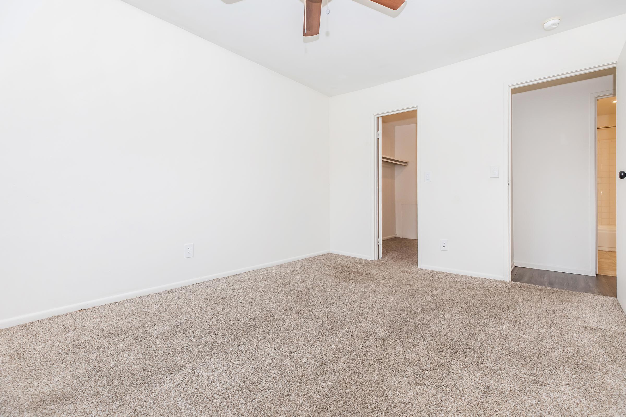 A small, empty room with light beige carpet and white walls. There is a ceiling fan and two doorways leading to separate spaces, one possibly a closet. Natural light might be coming from an unseen window, creating a bright and airy feel in the otherwise unadorned space.