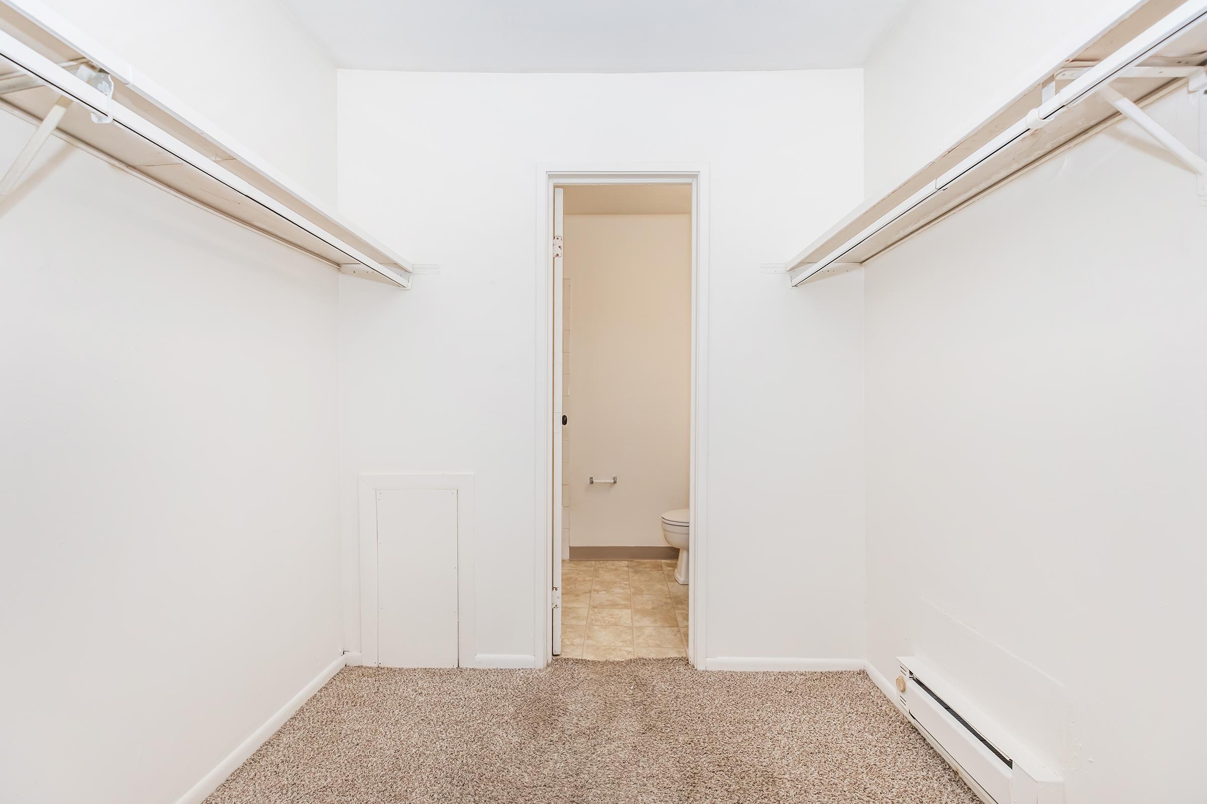 A spacious, empty closet with light beige carpet and white walls. The closet features two hanging rods on either side. A door leads to a small, visible bathroom area in the back. The overall atmosphere is bright and minimalist, providing ample storage space.