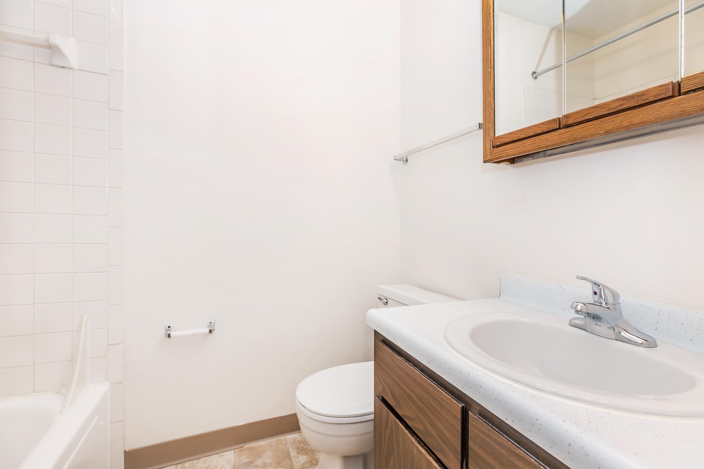 A clean, empty bathroom featuring a white toilet, a sink with a countertop, and a mirror above the sink. The walls are tiled in light colors, and there is a towel bar. A bathtub is visible in the background, and the floor has a light, textured finish.