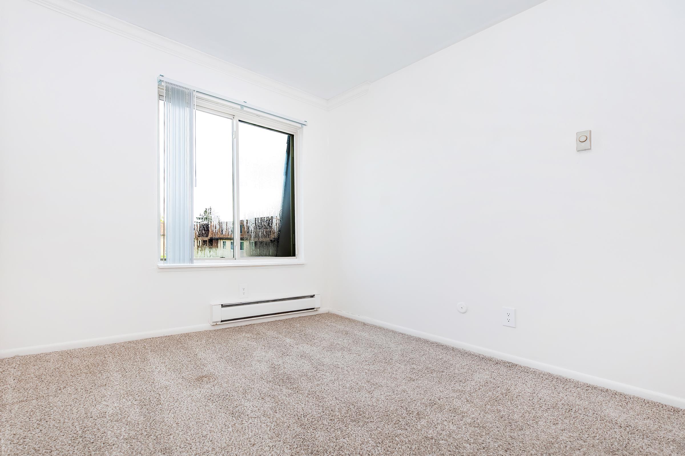 A bright and empty room with light-colored walls and a carpeted floor. There is a large window on one wall with sheer blinds, allowing natural light to enter. The room has minimal furnishings and a heating unit on the wall, creating a spacious and clean atmosphere.