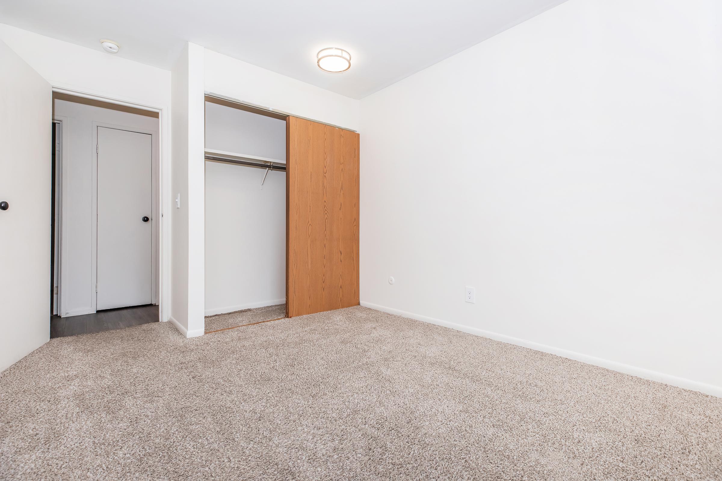 A clean, empty room with light-colored walls and a carpeted floor. There's a closet with wooden doors on one side, and a white door leading to another area. Natural light illuminates the space from above, creating a bright and airy atmosphere.