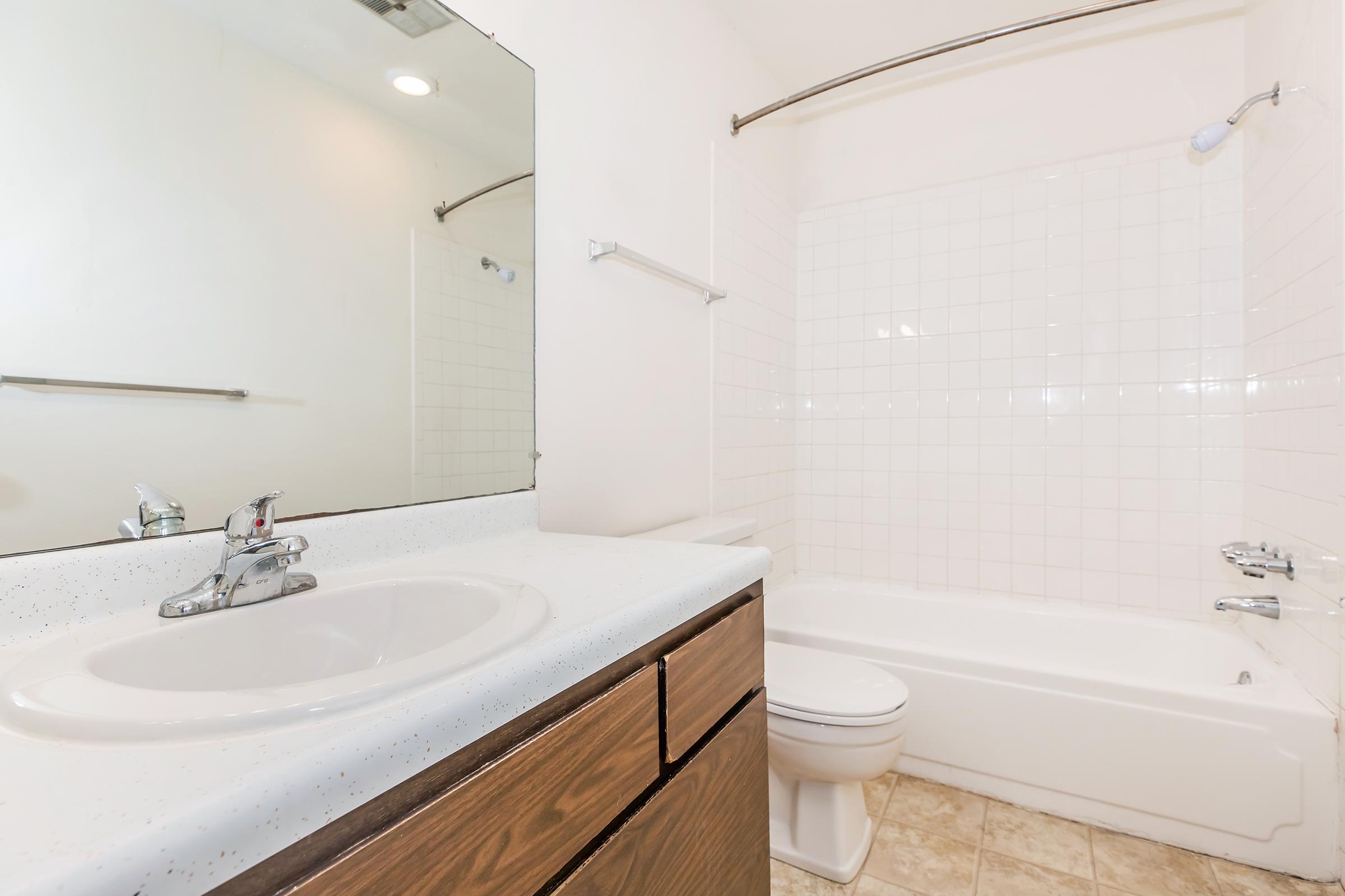 A clean and modern bathroom featuring a white bathtub with a shower, a mirror above a countertop sink, and wooden cabinetry. The walls are tiled in white, and the flooring is a light beige tile, creating a bright and spacious atmosphere.
