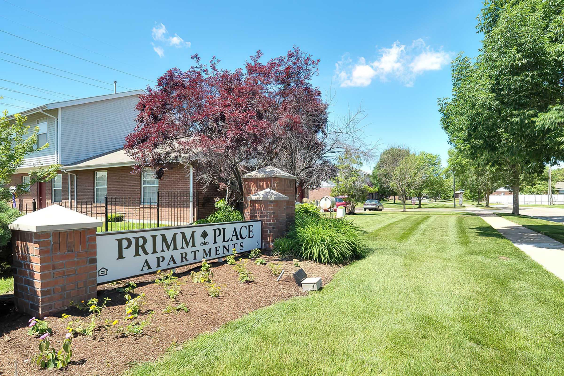 A well-maintained sign for Primm Place Apartments is surrounded by green grass and colorful landscaping. The backdrop features a sunny blue sky with a few clouds, and there are trees lining the walkway, creating a pleasant and inviting atmosphere.
