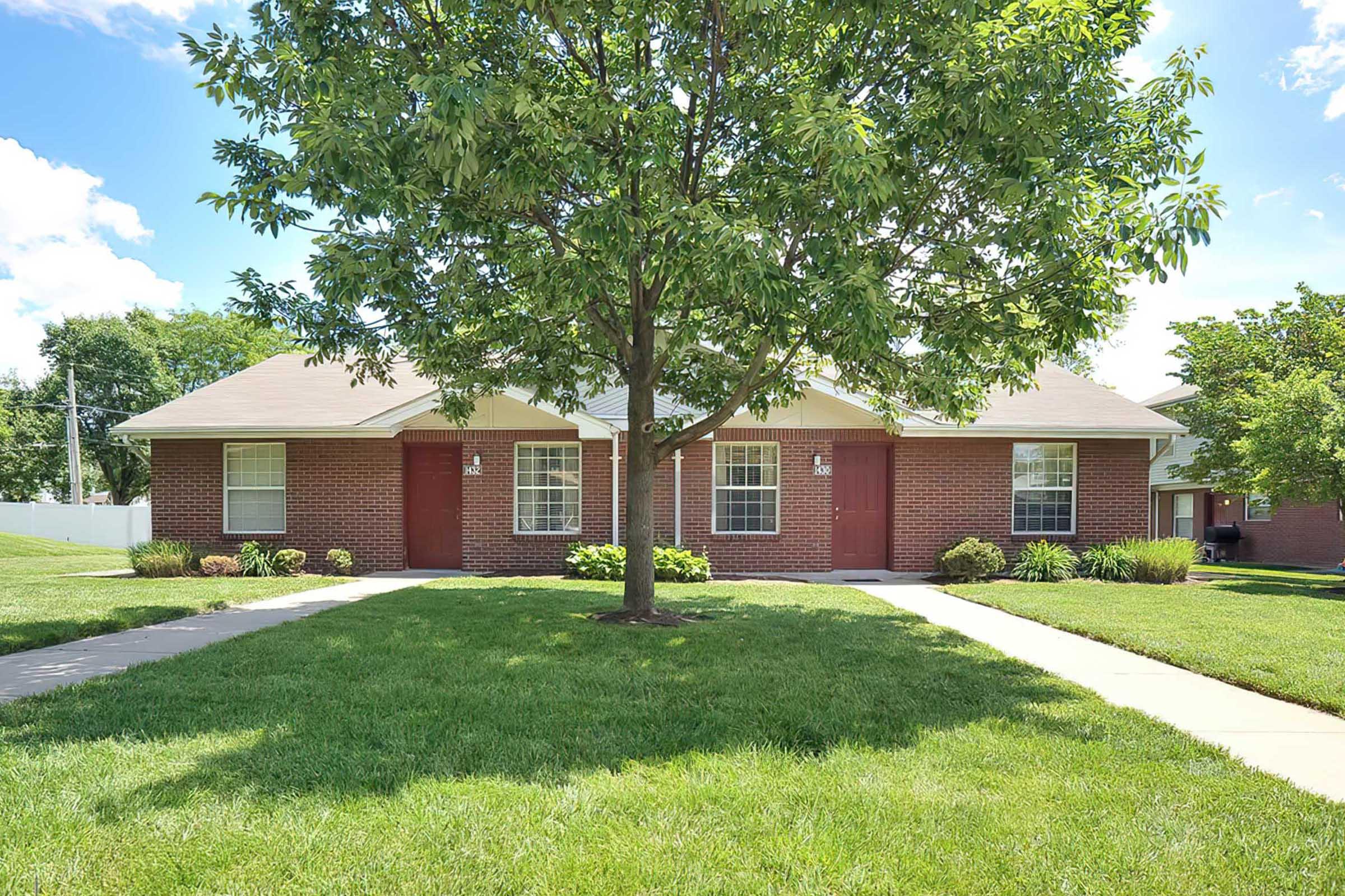 A view of a brick duplex, featuring two front doors, framed by a lush green lawn and a large tree in the center. The sky is clear with a few clouds, and there are shrubs and plants along the walkway leading to the entrances.