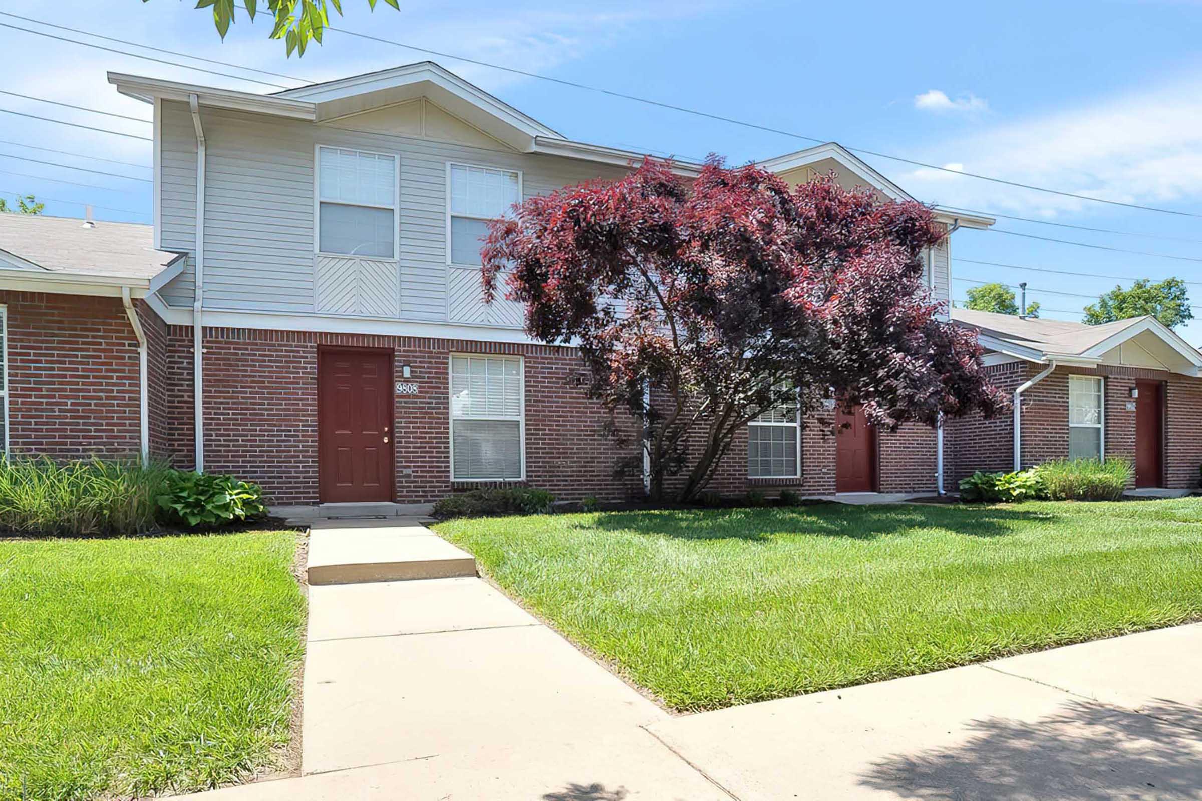 A two-story residential building with a brick facade surrounded by a well-maintained lawn. In front, there are several small shrubs and a tree. The sky is clear and blue, suggesting a sunny day. The pathway leads to the entrance of the building.