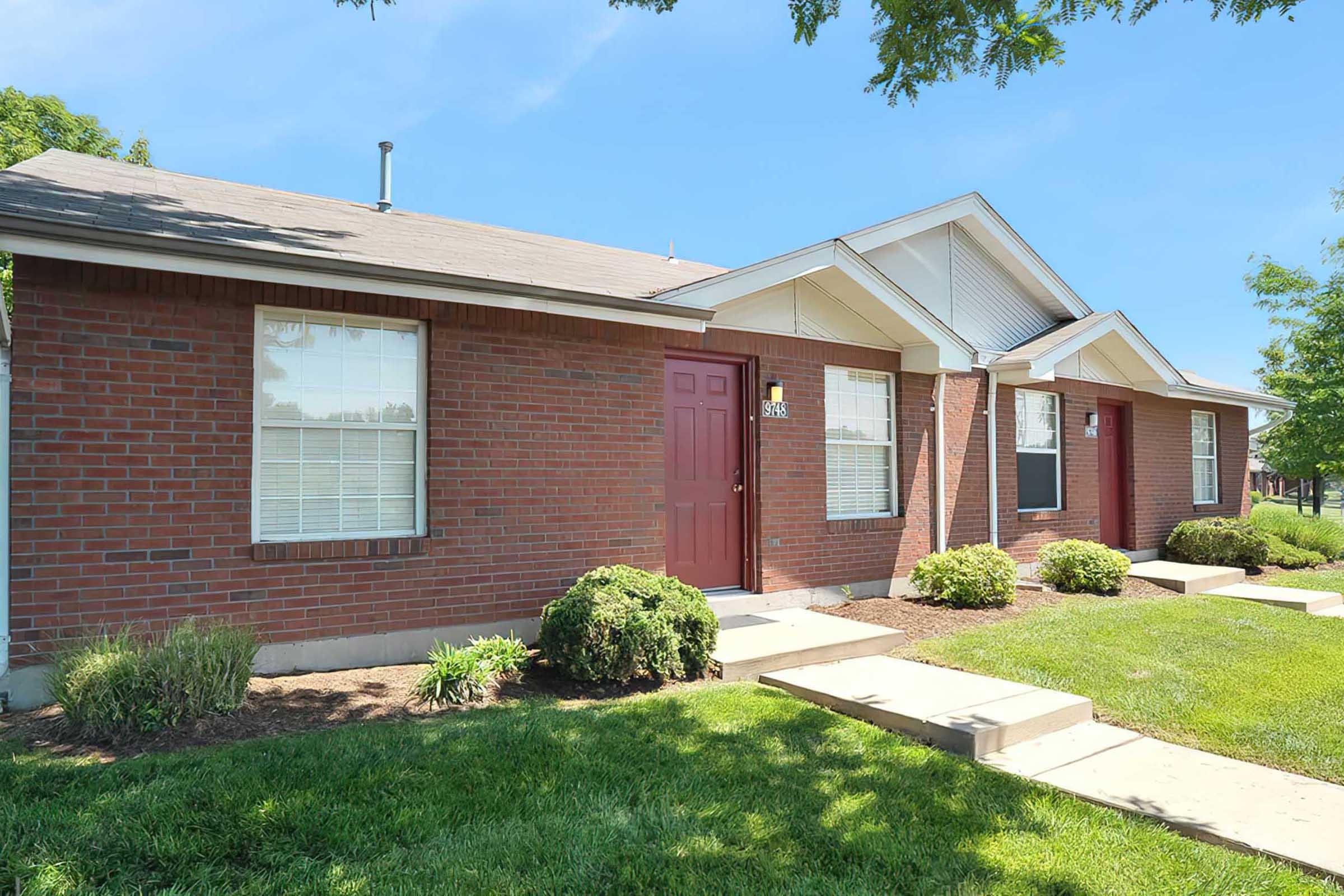 A row of brick houses with red doors and white trim under a clear blue sky. The front yards feature well-maintained grass and small shrubs. The buildings appear to be part of a residential neighborhood.