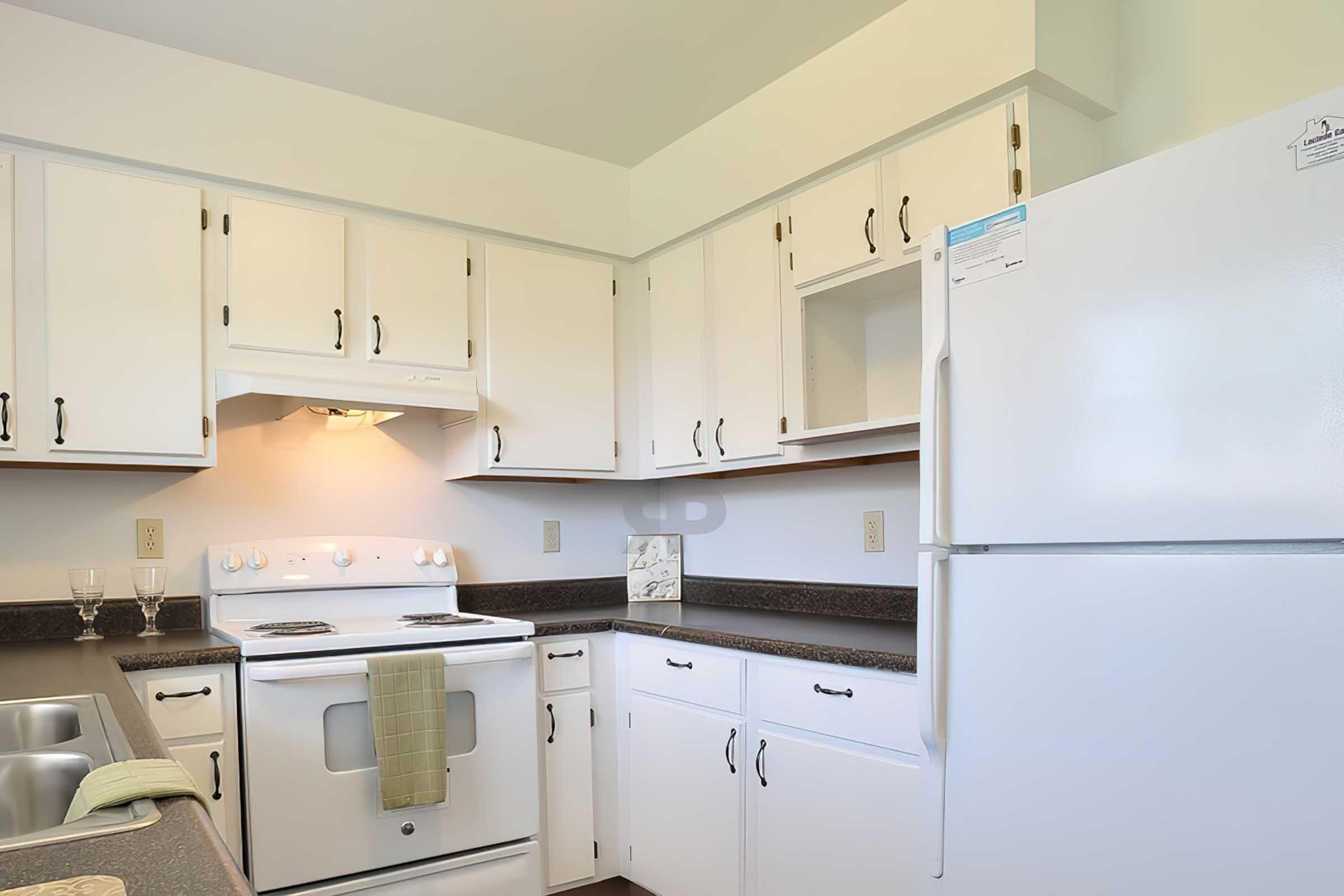 A clean and modern kitchen featuring white cabinetry, a white refrigerator, and a stove. The countertop is dark, and there are two sinks. A light fixture illuminates the space, and a green dish towel is hung on the stove. The overall atmosphere is bright and inviting.