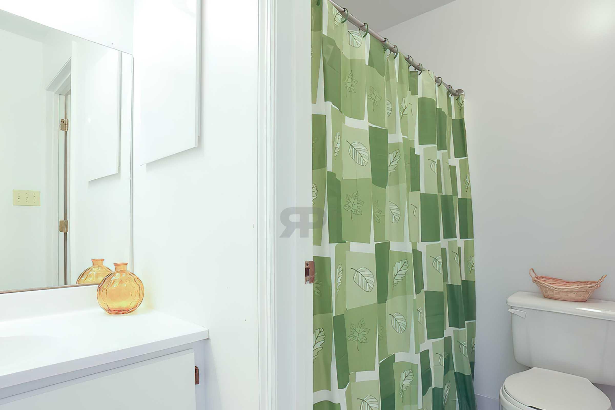 A bathroom featuring a green and white patterned shower curtain, a white toilet, and a simple vanity with a mirror. Two decorative orange vases sit on the vanity. The overall color scheme is light and fresh, contributing to a clean and inviting atmosphere.