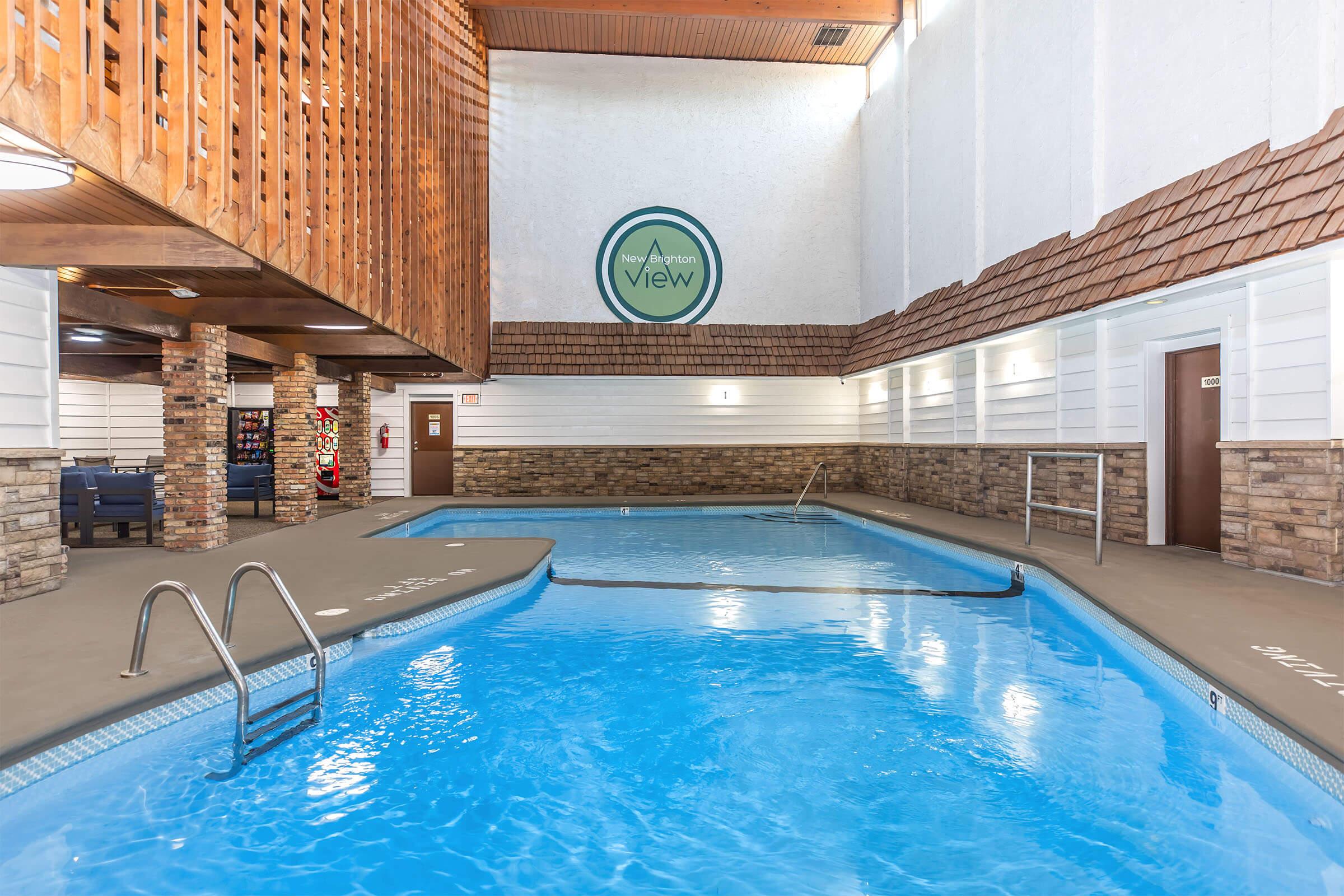Indoor swimming pool area with two connected pools. The first pool features a ladder, while the second is a smaller round pool. Walls are made of stone and wood accents, with a small area for seating. A circular logo on the wall reads "View." Natural light fills the space from overhead.