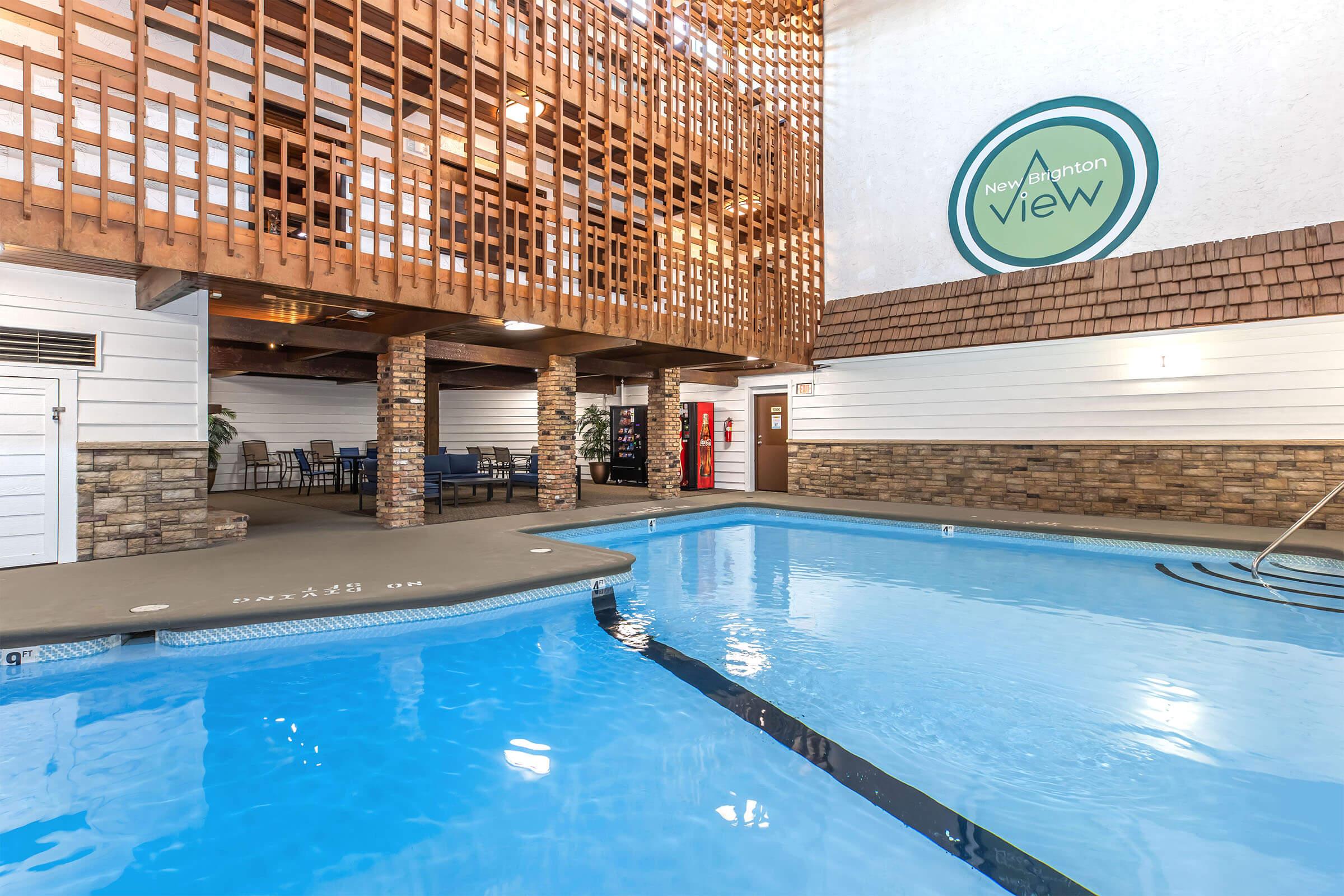 Indoor swimming pool area featuring clear blue water, surrounded by stone walls and wooden accents. There are lounge chairs and a dining area visible beyond the pool, with a sign reading "New Hamilton View" on the wall. Natural light filters through a wooden structure overhead.