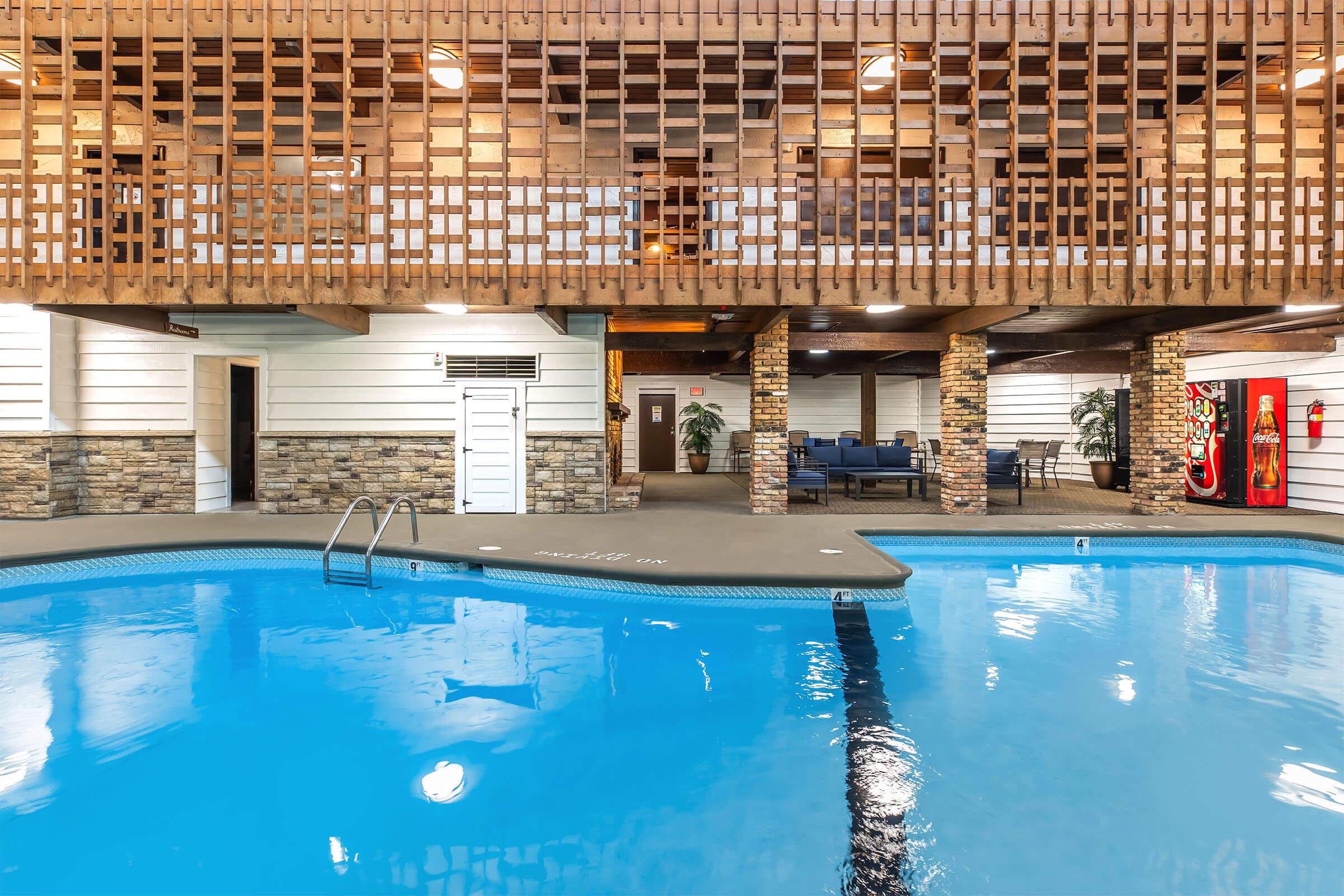 Indoor swimming pool with clear blue water, surrounded by a deck area featuring wooden paneling and stone accents. There are lounge chairs and a cozy seating arrangement in the background, along with indoor plants and arcade games visible on the side.