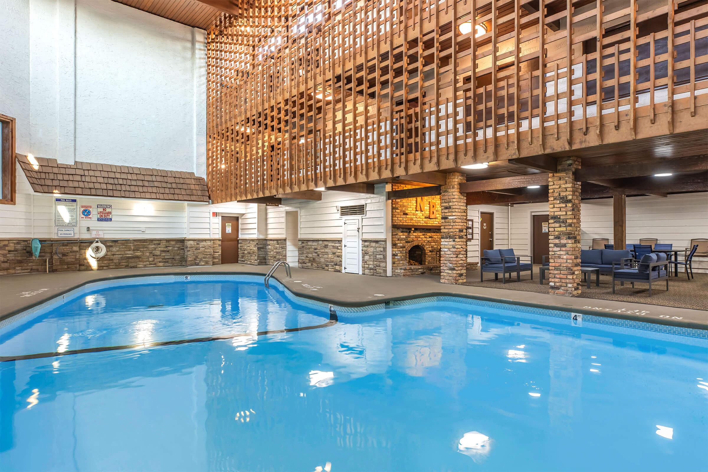 Indoor swimming pool area with clear blue water, surrounded by stone pillars and wooden accents. Comfortable seating is visible along the poolside, and a balcony overlooks the space. Natural light filters through the ceiling, creating an inviting atmosphere.