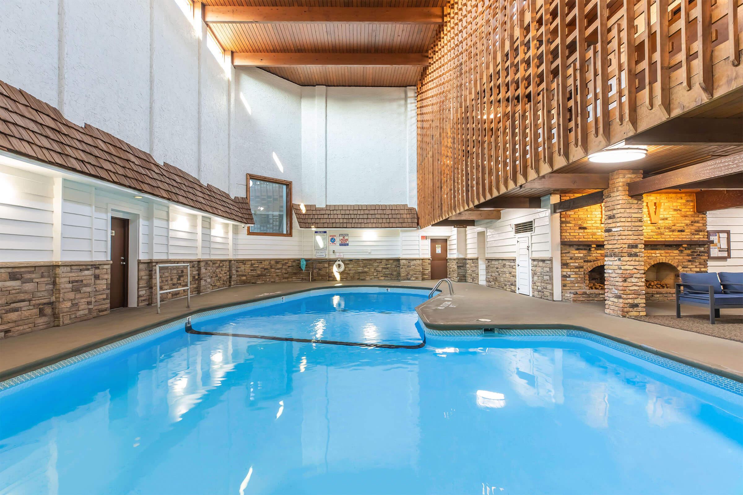 Indoor swimming pool with a clear blue water surface. The area features a stone wall and wooden accents, with large windows allowing natural light to illuminate the space. There are lounge chairs near the pool, and a fireplace is visible in the background, creating a cozy atmosphere.