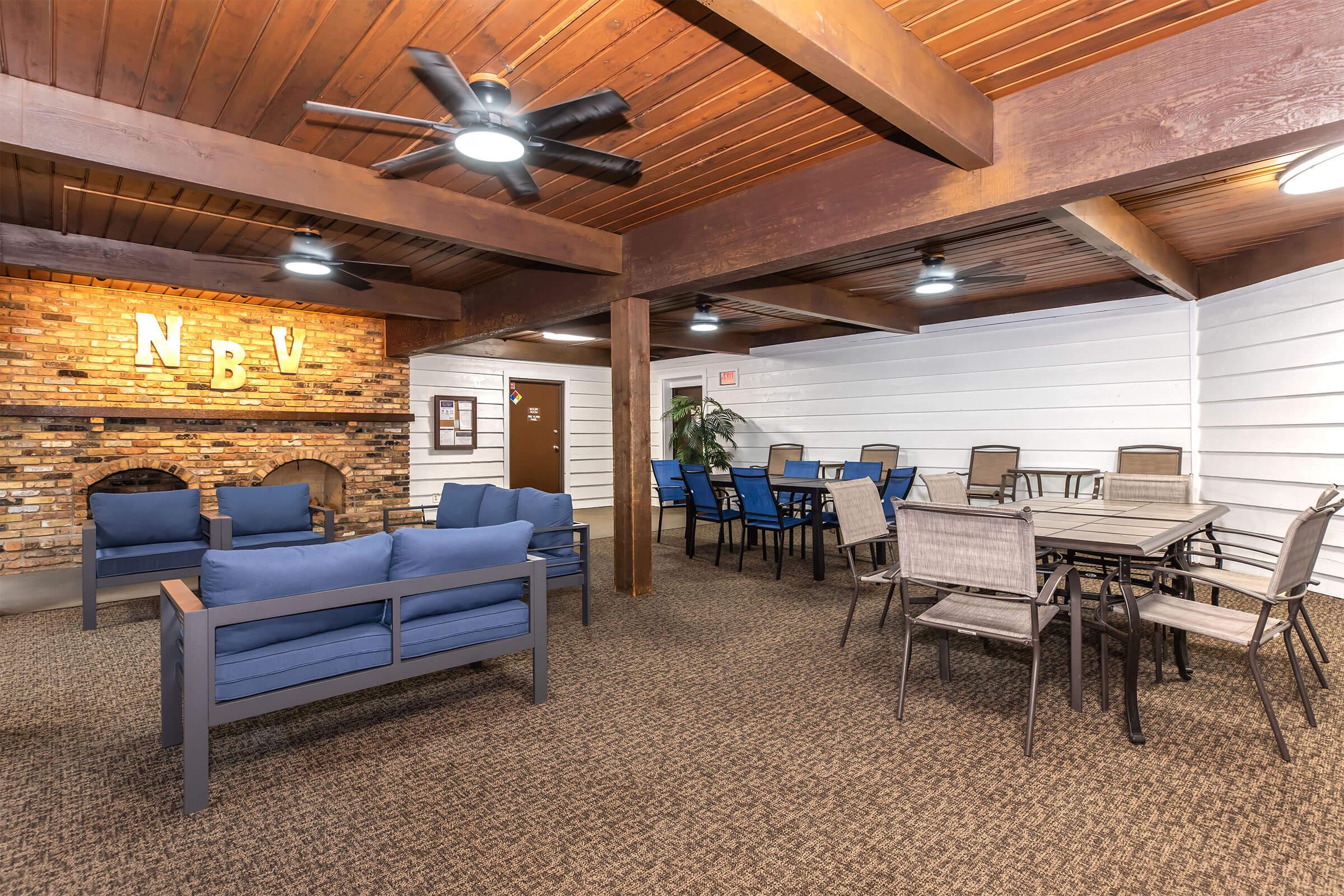 A cozy indoor lounge area featuring a brick fireplace with "NBV" displayed, comfortable blue sofas, and a variety of dining tables and chairs. The space is well-lit, with wooden beams across the ceiling and a welcoming atmosphere, perfect for social gatherings or relaxation.