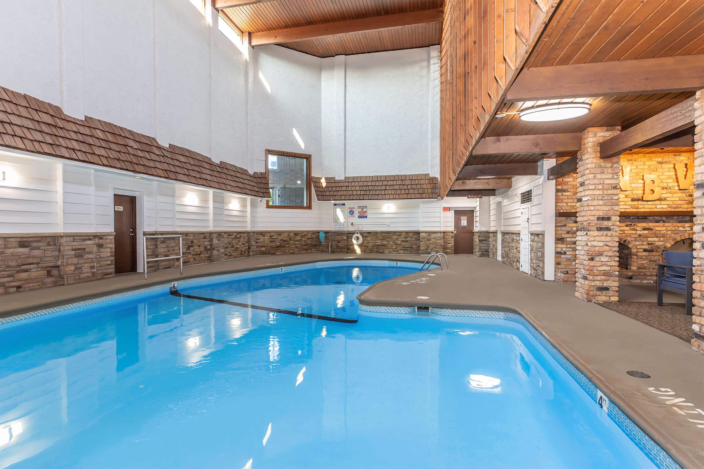 Indoor swimming pool with clear blue water, surrounded by wooden beams and stone accents. Sunlight streams through windows, illuminating the area. There are lounge chairs nearby and a pathway leading around the pool. The space has a welcoming and relaxed atmosphere, ideal for leisure activities.