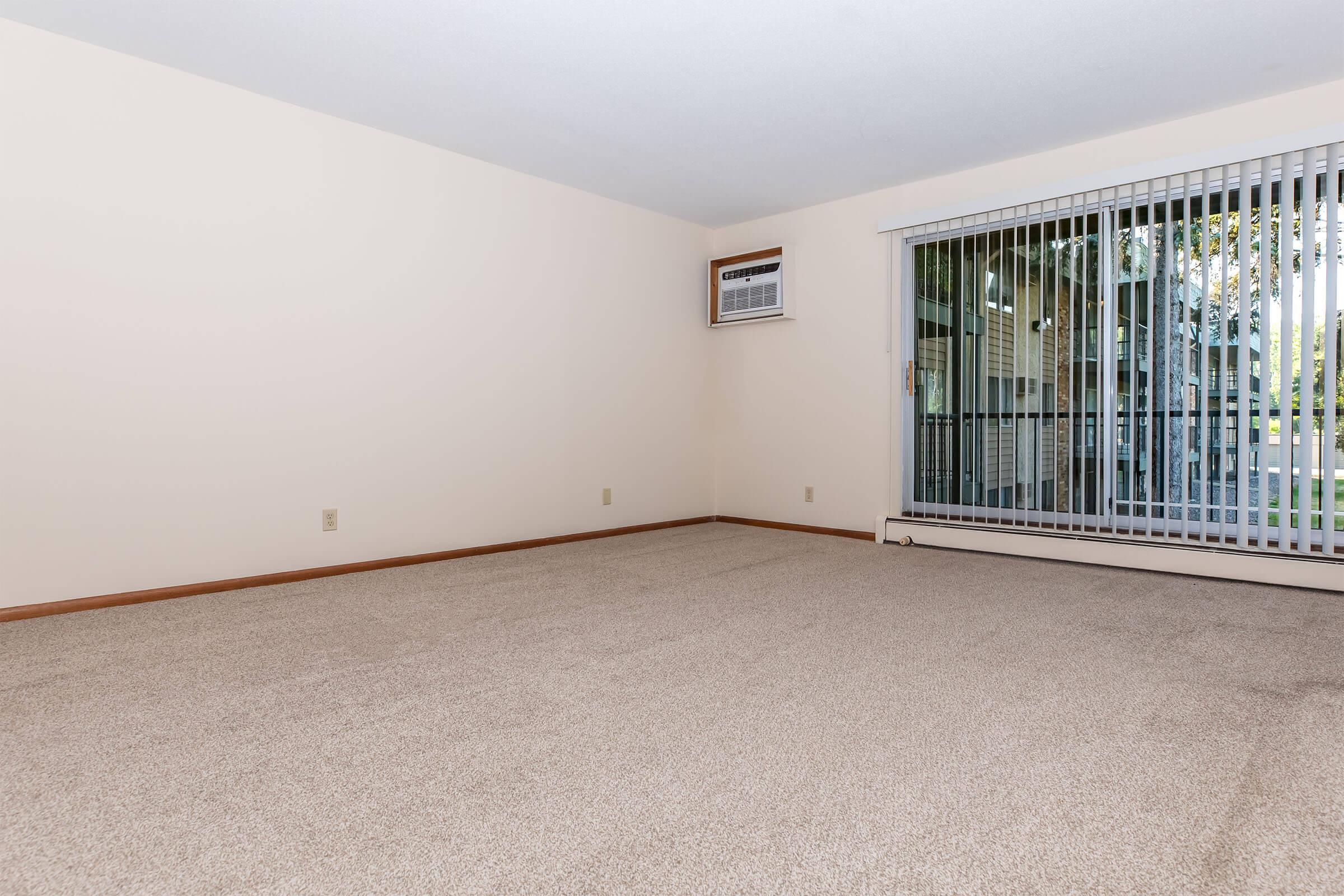 A spacious, empty room with light beige walls and carpet. One wall has a window with vertical blinds, and an air conditioning unit is mounted above it. The area has a clean, uncluttered look, making it feel bright and airy, perfect for various interior design possibilities.