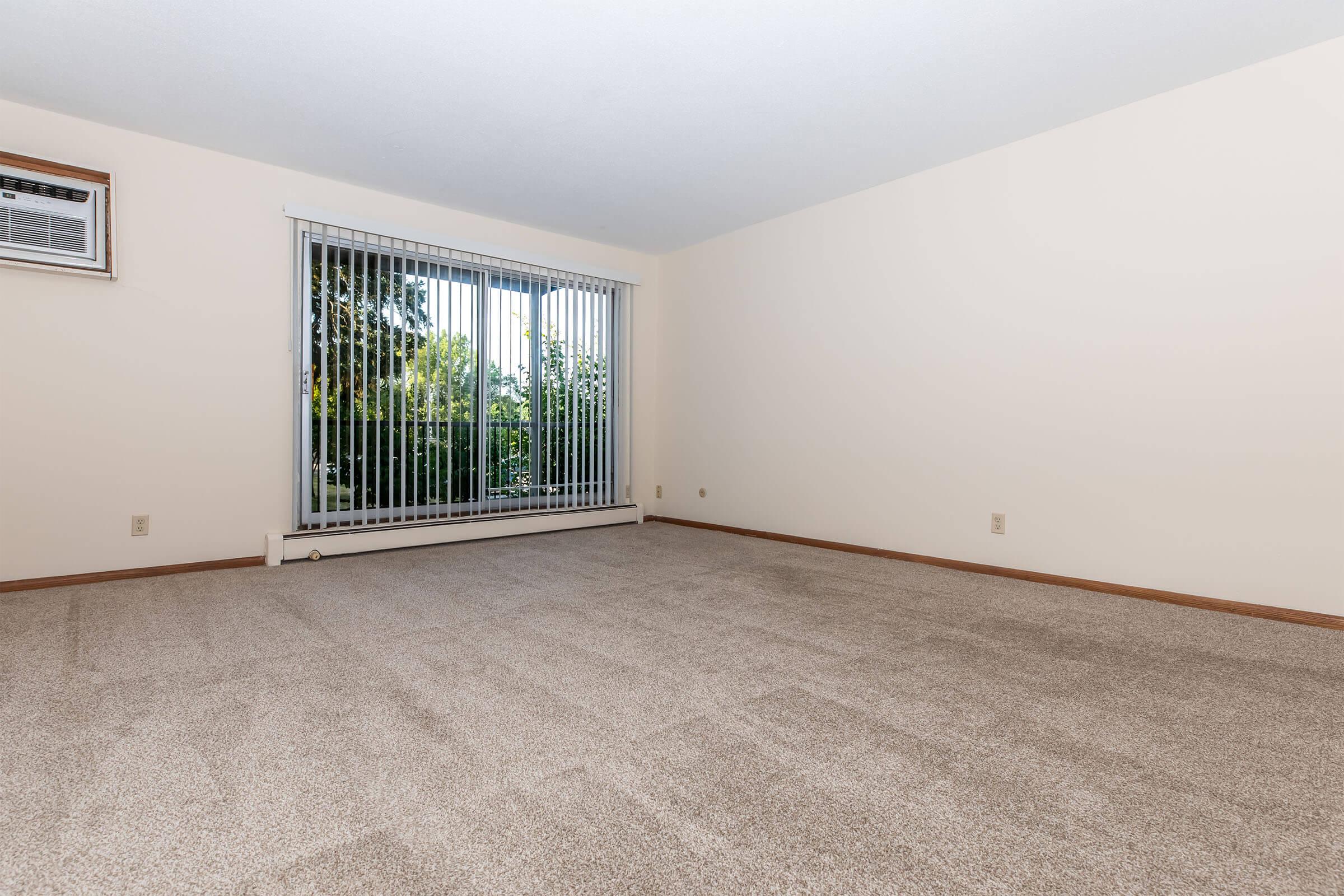 A spacious, empty room with light beige walls and a large window covered by vertical blinds. The floor is carpeted in a light shade, and there is an air conditioning unit on the wall. The window offers a view of greenery outside, indicating a bright and airy environment.