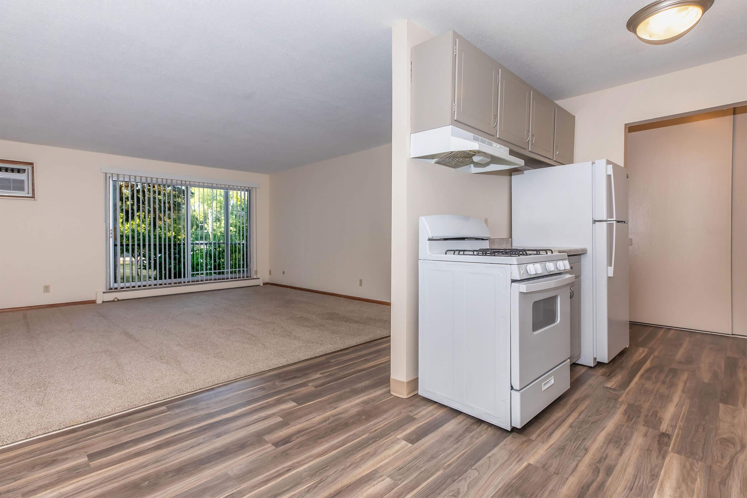 A modern kitchen with a white stove and refrigerator, featuring wood-like flooring. The adjacent living area has beige carpet and a large window with vertical blinds, allowing natural light. The walls are painted in a light color, creating a spacious and inviting atmosphere.