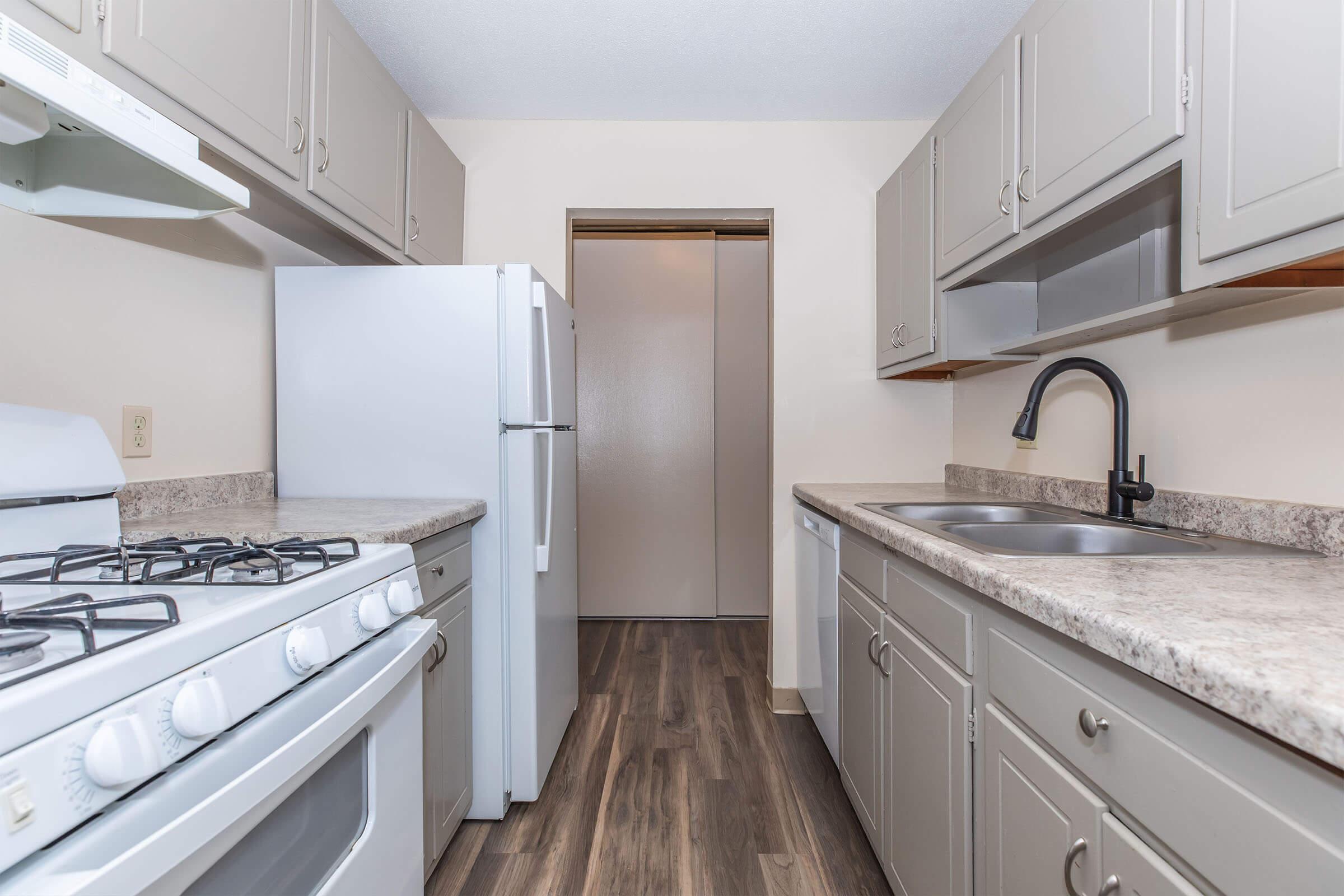 A modern kitchen featuring light gray cabinets, a gas stove, and a double sink. The countertops are a dark speckled design, and there are white appliances, including a refrigerator and a microwave. The room has a neutral-colored wall and a doorway leading to another room at the end of the space.