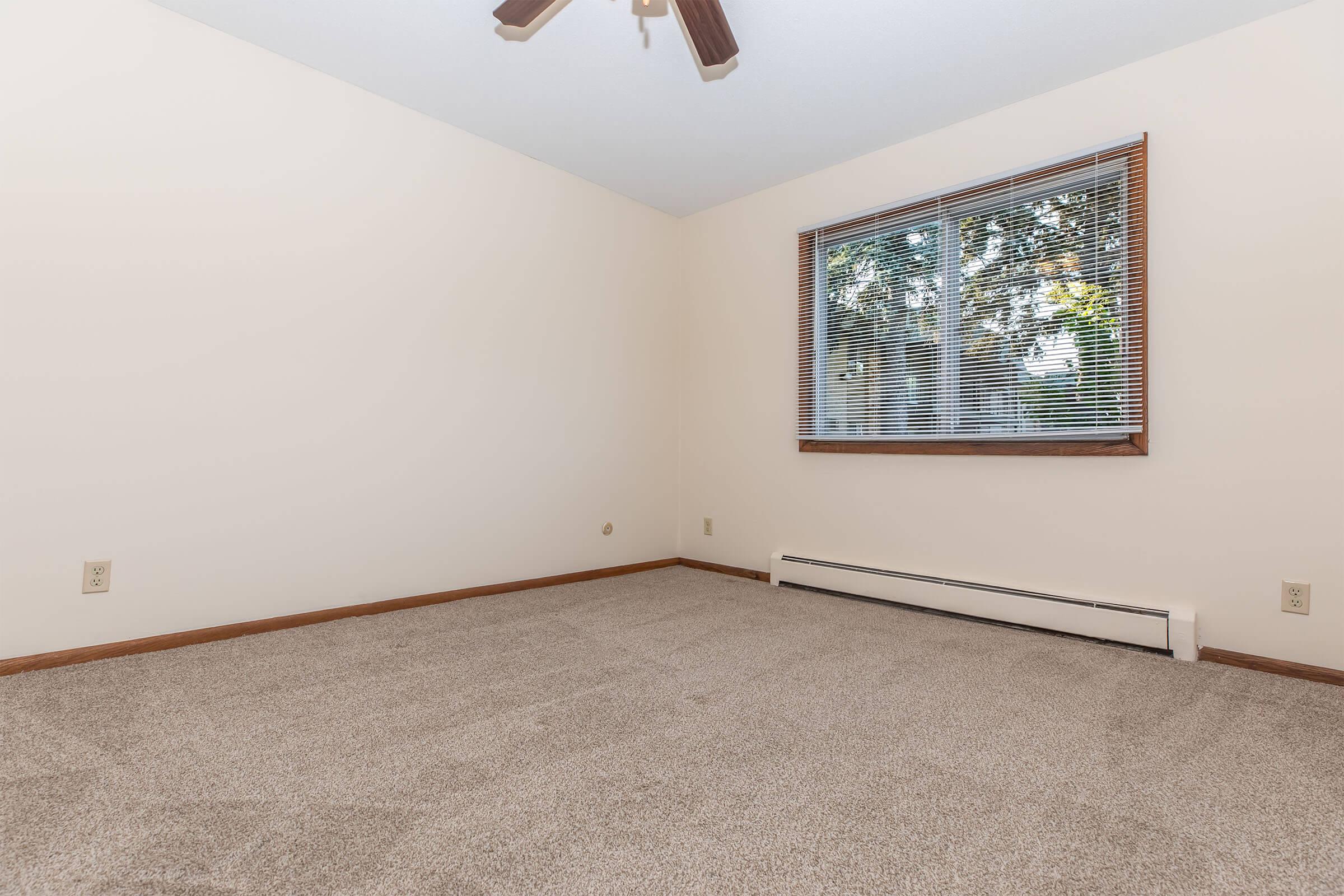 A vacant room with beige walls and a light brown carpet. A large window with blinds lets in natural light, showing greenery outside. The ceiling features a ceiling fan, and there are baseboard heaters along the walls. The room appears clean and uncluttered, ideal for various uses.
