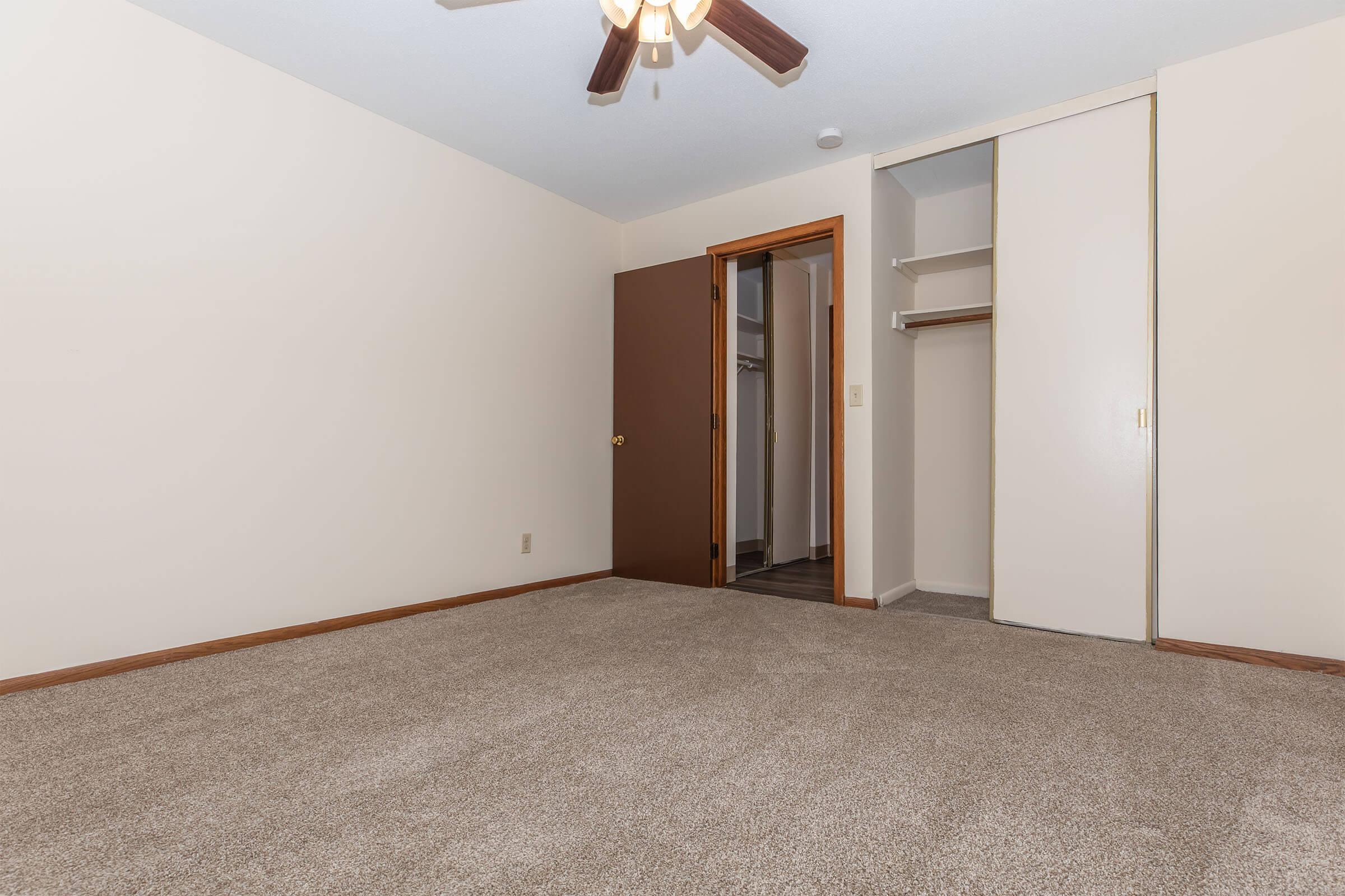 An empty bedroom with a ceiling fan and beige walls. The floor is covered in light brown carpet. There is a closed closet door on the left and an open closet space visible behind it. There are no furniture or decorations, creating a spacious and clean appearance.
