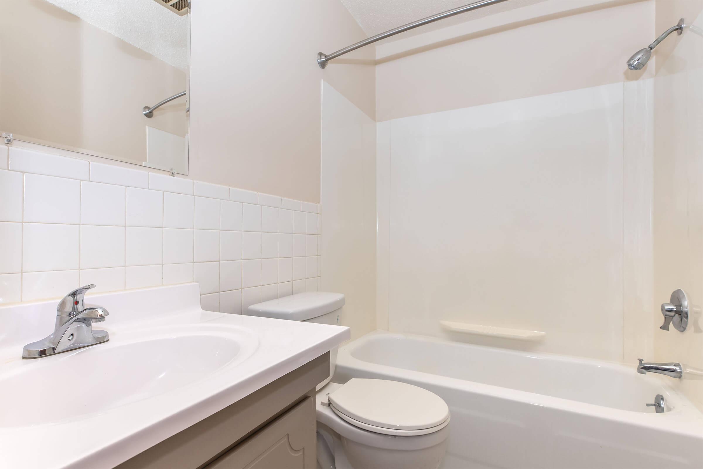 A clean, modern bathroom featuring a white bathtub with an attached shower, a toilet, and a vanity with a sink. The walls are painted light beige, and the tile backsplash contrasts with the neutral tones. Natural light illuminates the space, highlighting its tidy and spacious layout.