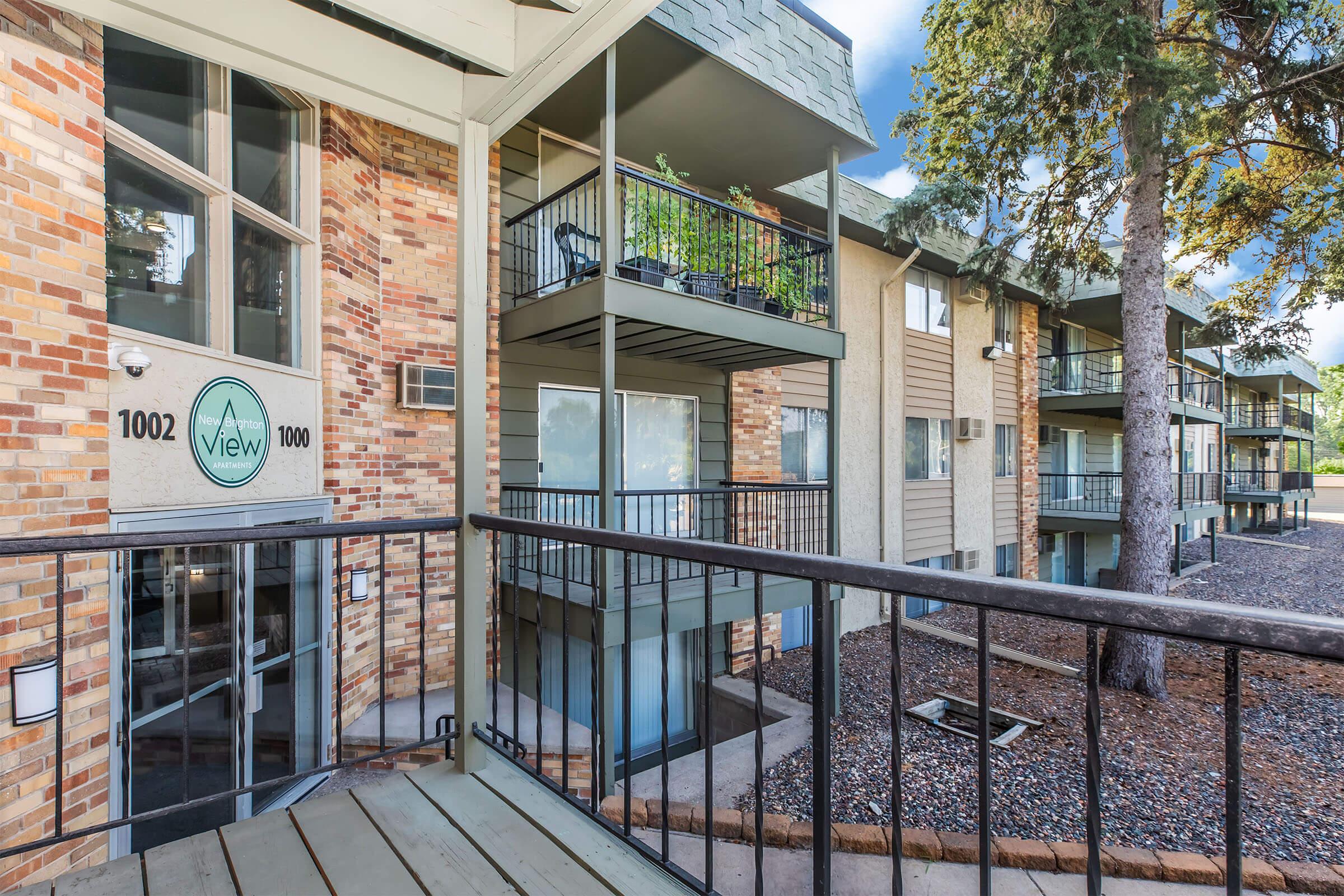 View of an apartment complex exterior, featuring multiple beige and brick buildings with balconies. The entrance displays the building number 1000 and the name "View." Surrounding the complex are trees and landscaped areas with gravel paths. Bright blue sky above.