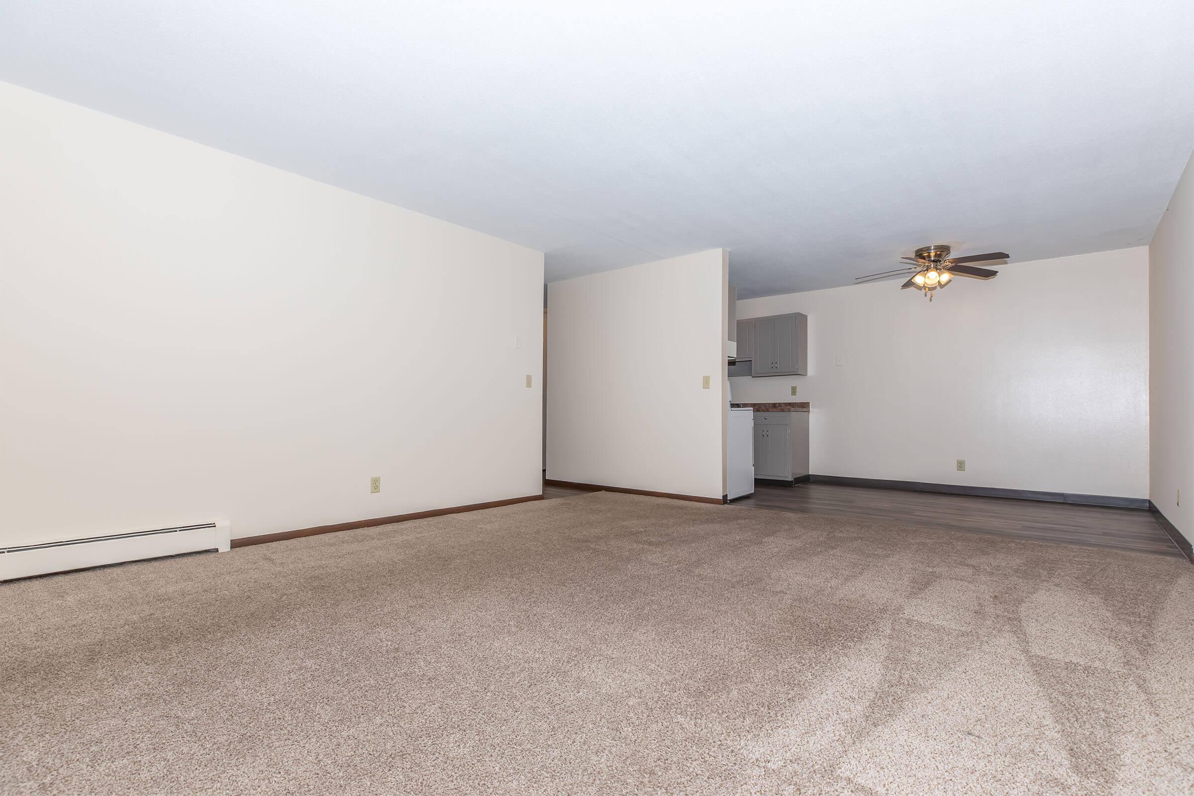 A spacious, empty living area with beige carpet and light-colored walls. There is a ceiling fan in the corner and a partial view of a kitchenette in the background. The room has no furniture, creating a blank canvas for potential furnishings and decorations.