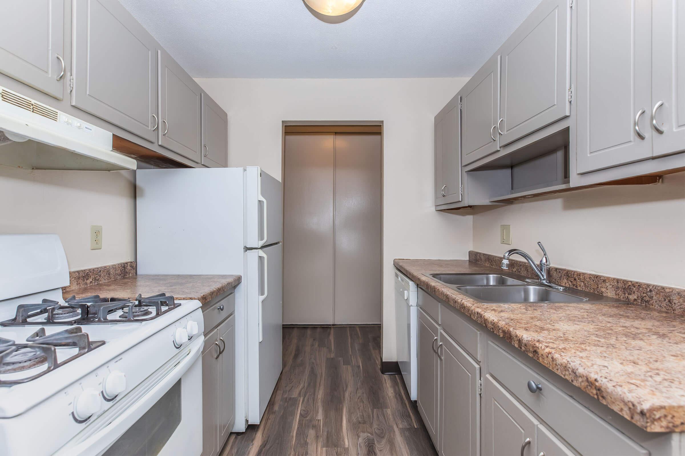 A clean, compact kitchen featuring light gray cabinets and countertops, a white gas stove, a refrigerator, and a double-basin sink. The kitchen has wooden flooring and a doorway leading to another room in the background. Natural light illuminates the space from a ceiling light fixture.